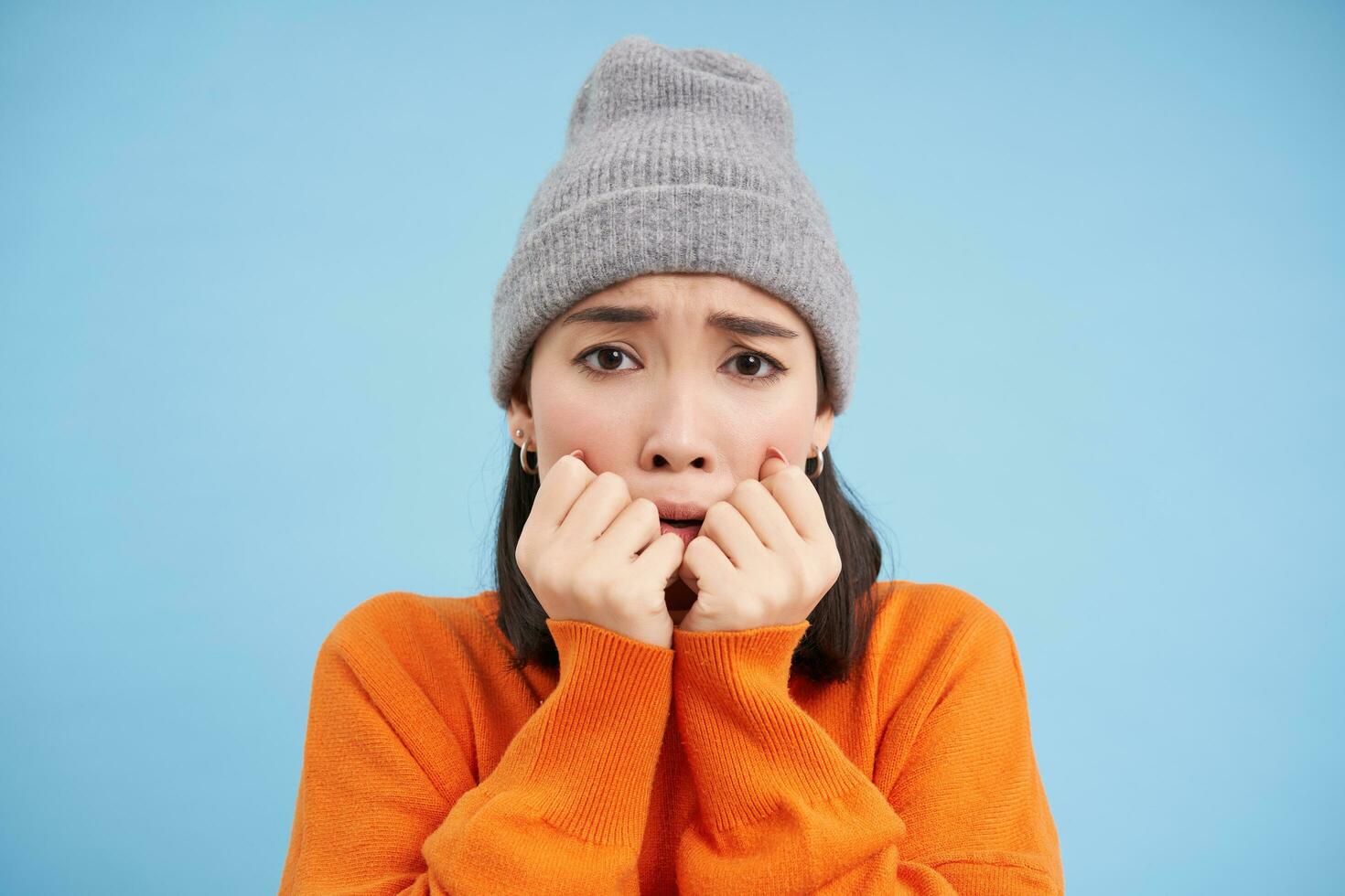 Portrait of asian girl in warm hat, looks with fear, shaking and trembling scared, standing frightened against blue background photo