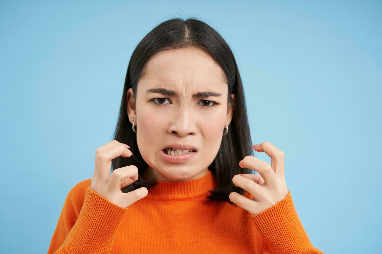 Close up of angry japanese woman, clenches her fists and looks furious, outraged and annoyed, stands over blue background photo