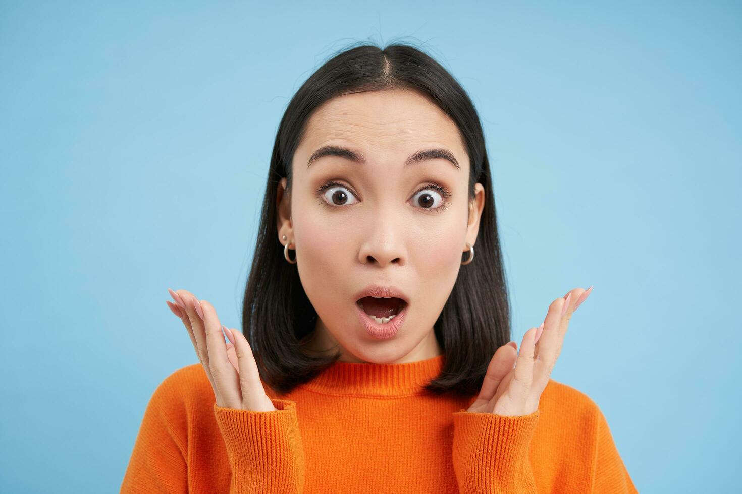 Close up portrait of amazed brunette girl says WOW, amazed by smth. Asian woman looks surpirsed at camera, impressed reaction, blue background photo