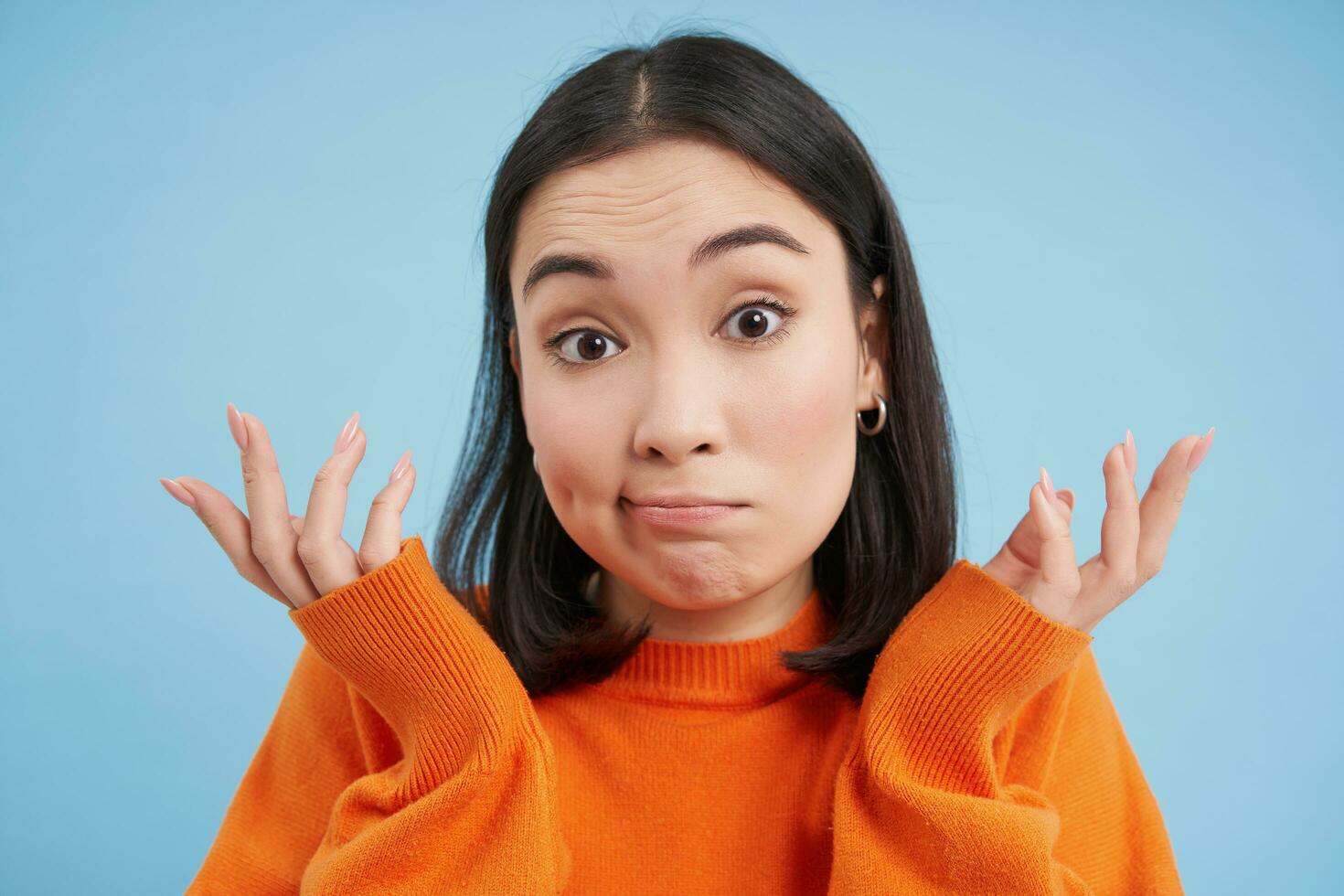 Close up of skeptical japanese girl, shrugs shoulders and looks confused, puzzled, stands over blue studio background photo