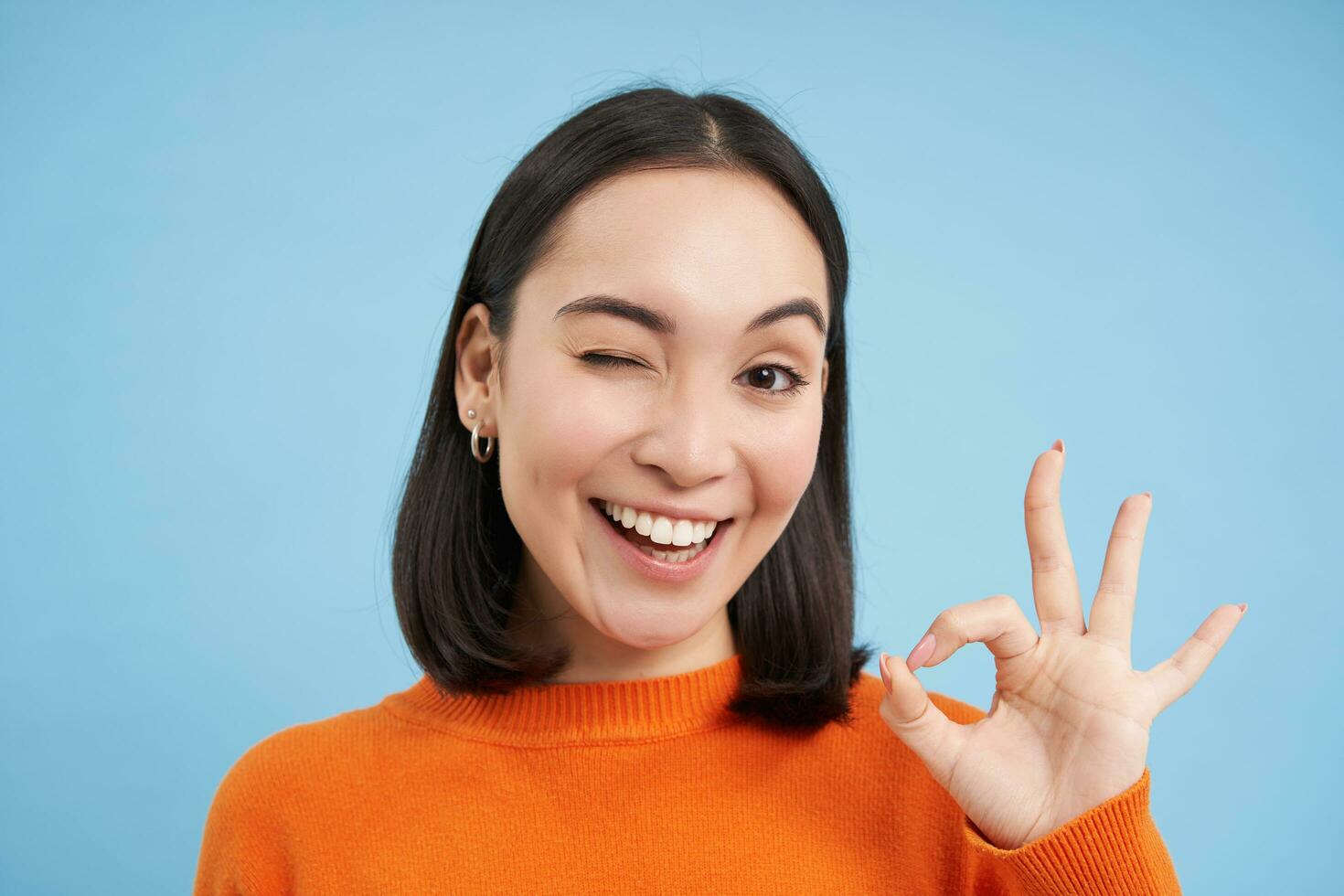 cerca arriba retrato de sonriente hermosa asiático mujer, muestra bueno, Okay bien firmar, aprueba algo, aprueba, da positivo comentario, azul antecedentes foto