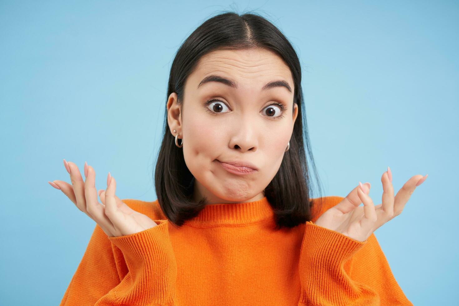 Close up of surprised japanese woman, shrugs and spreads hands sideways, looks confused, stands over blue background photo