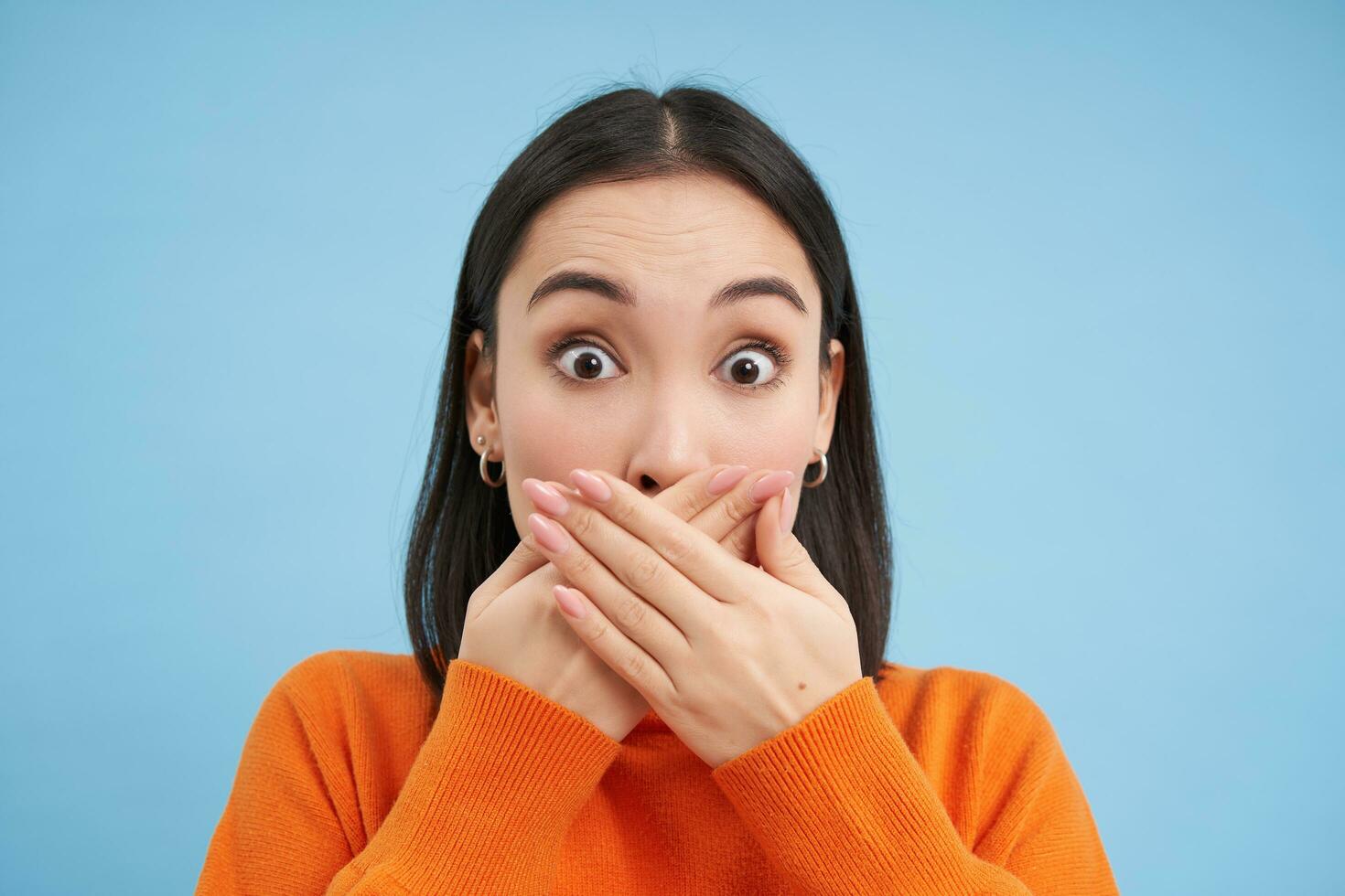 Close up portrait of shocked korean woman shuts her mouth with startled, impressed face expression, stands over blue background photo