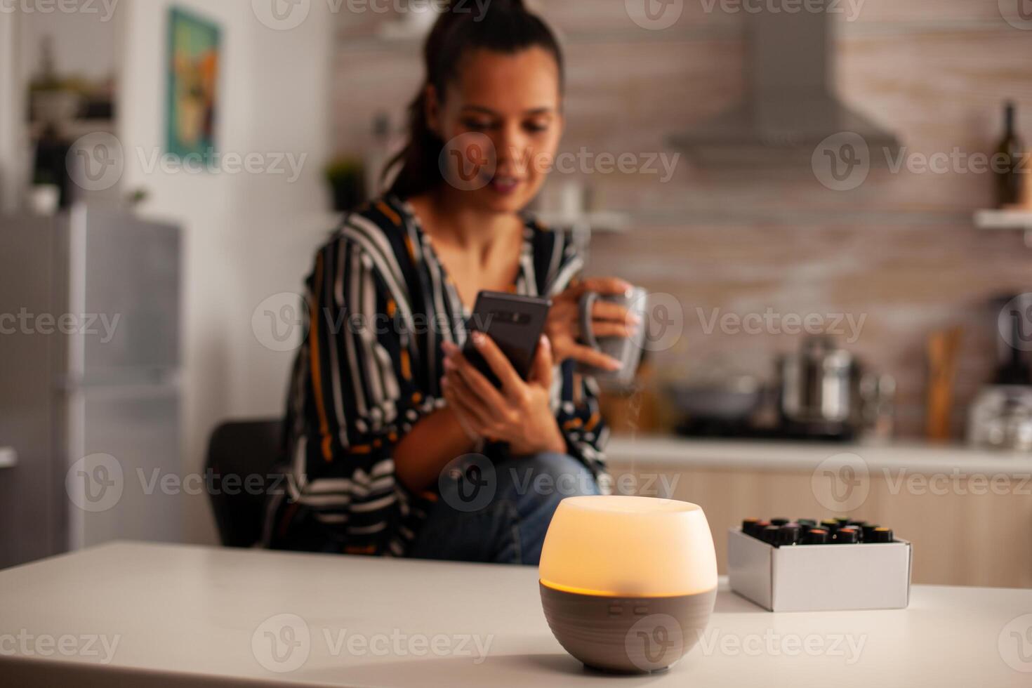 Woman watching video on phone and enjoying steam with essential oils from diffuser. Aroma health essence, welness aromatherapy home spa fragrance tranquil theraphy, therapeutic steam, mental health treatment photo