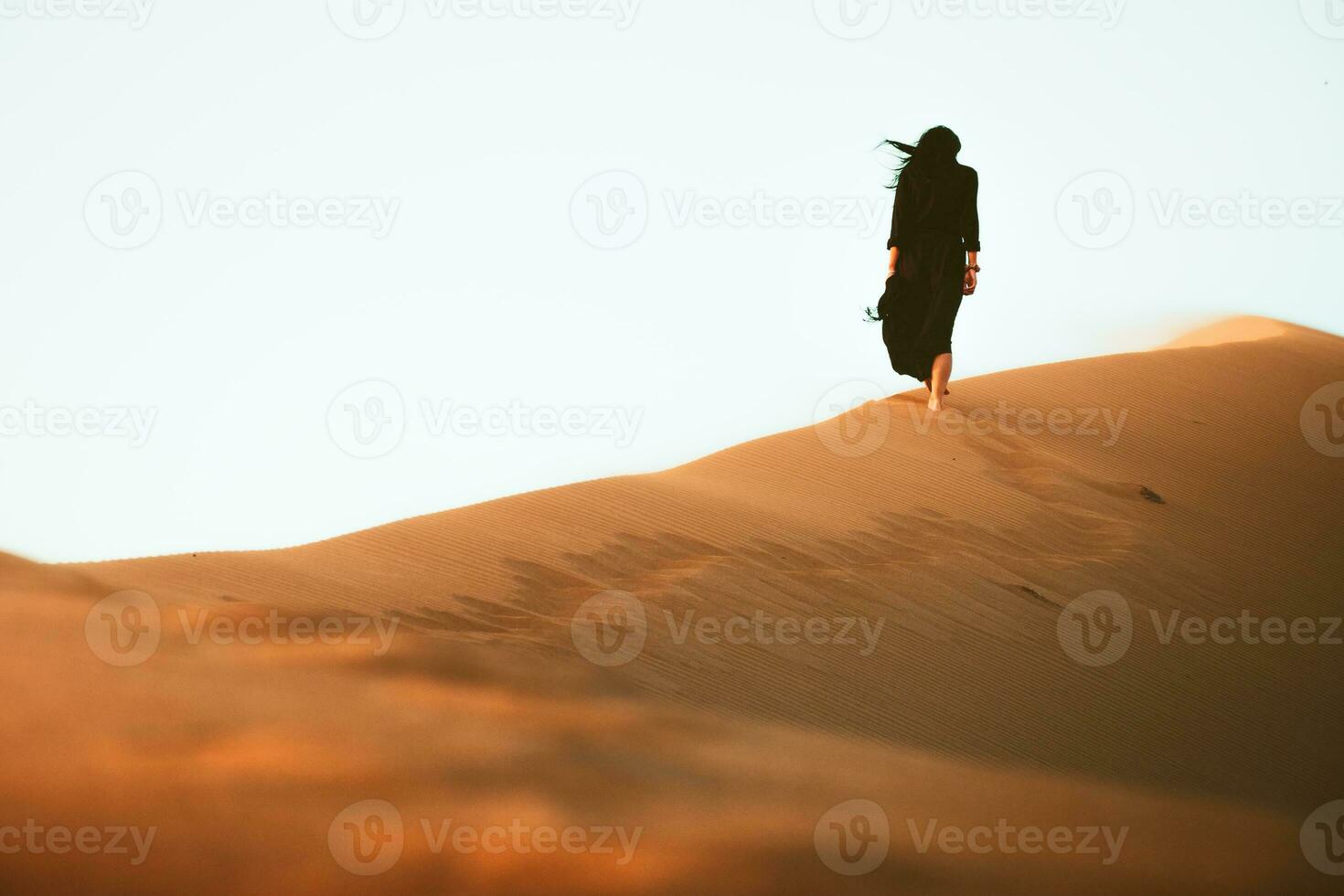 Low angle beautiful woman in long black dress barefoot walk up follow walk on golden Sahara desert dunes photo