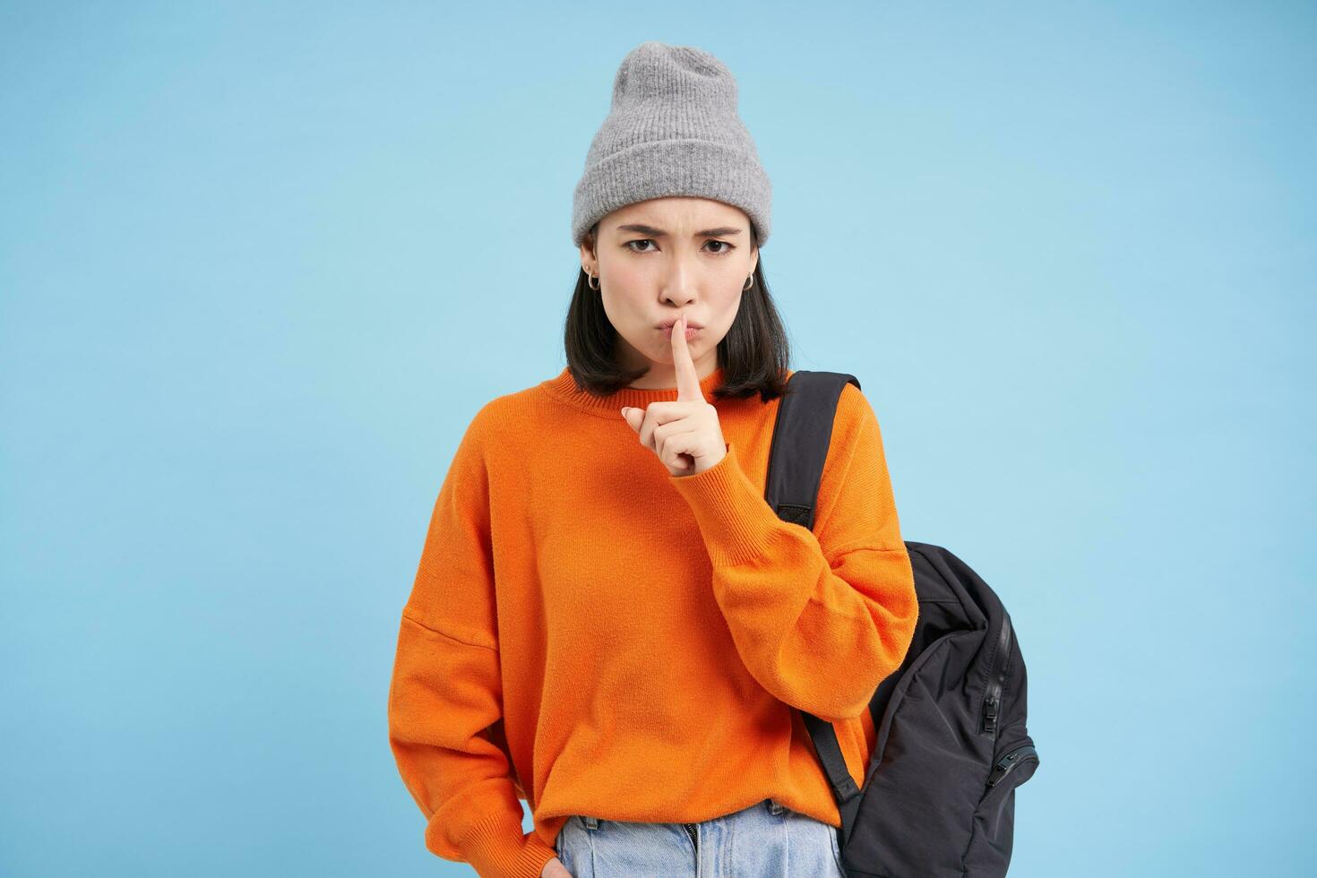 Asian girl shows shush, shh gesture, puts finger over lips, keep quiet sign, stands over blue background with backpack and wears hat photo