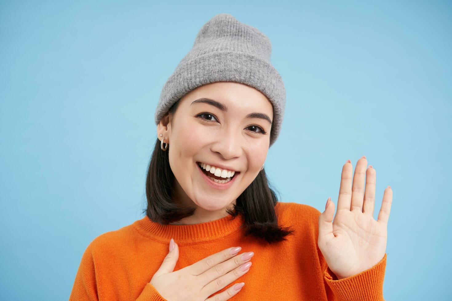 cerca arriba de linda coreano niña en gorro, levanta uno mano y presenta sí misma, saluda tú, sonrisas y mira amigable, azul antecedentes foto