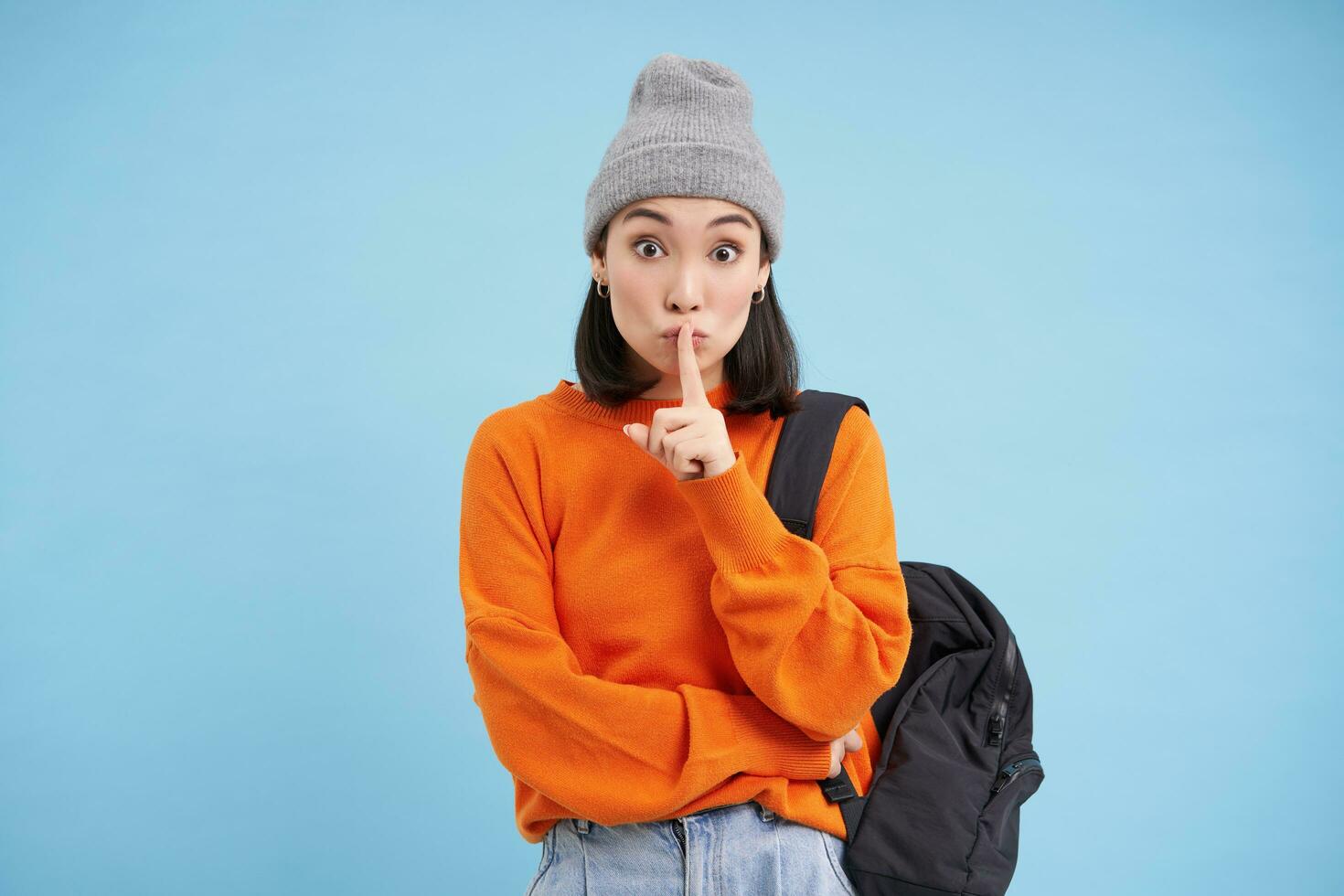 Asian girl shows shush, shh gesture, puts finger over lips, keep quiet sign, stands over blue background with backpack and wears hat photo