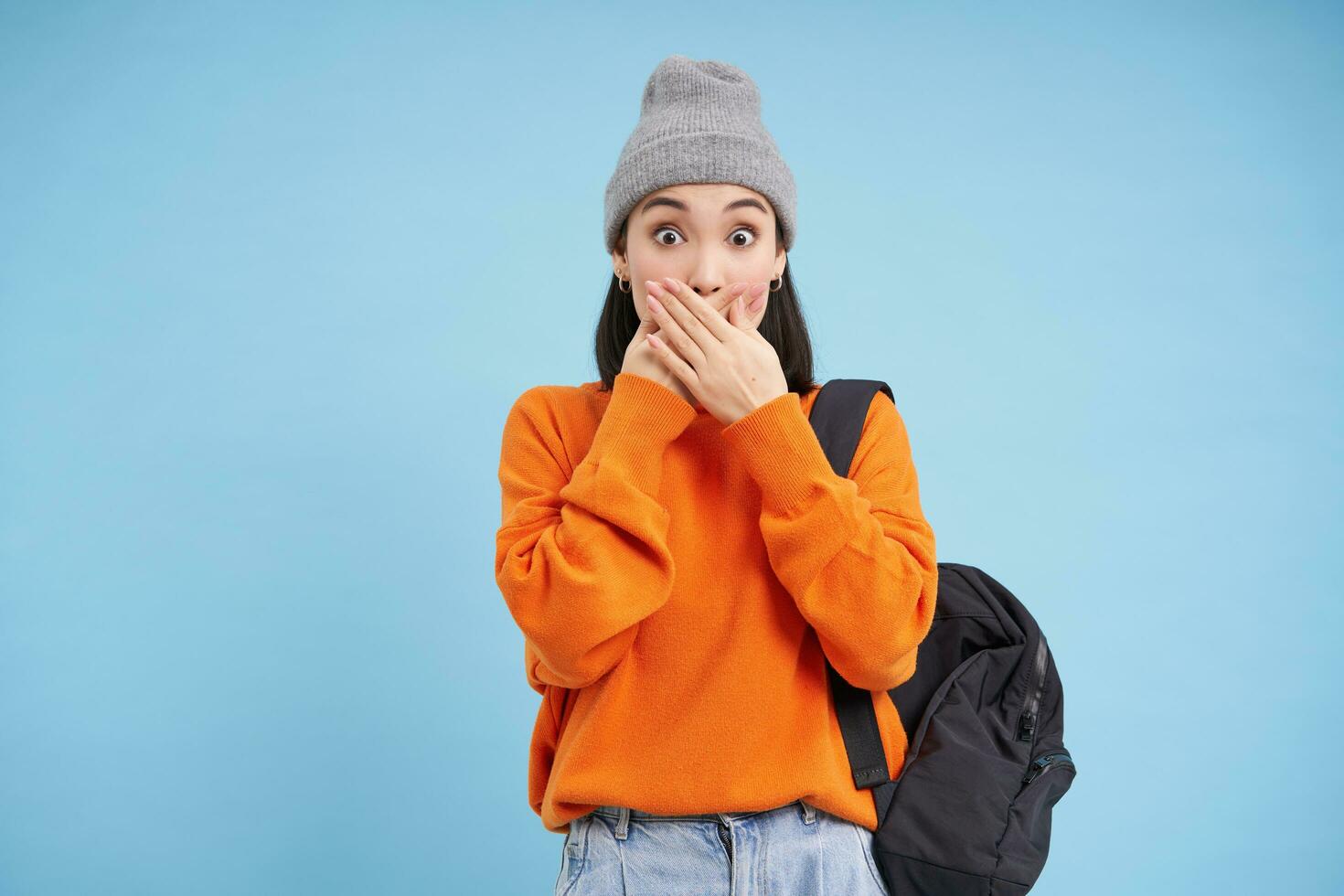 Portrait of korean girl in hat, covers her mouth with hands, looks shocked, amazed by gossips, stands over blue background photo