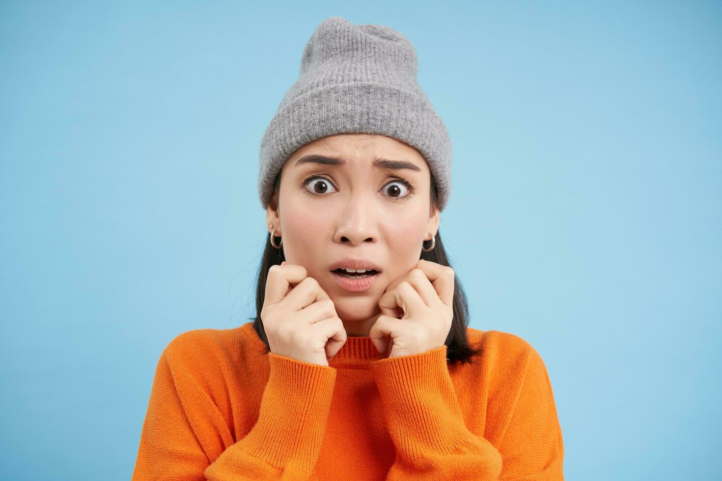 Portrait of asian girl in warm hat, looks with fear, shaking and trembling scared, standing frightened against blue background photo