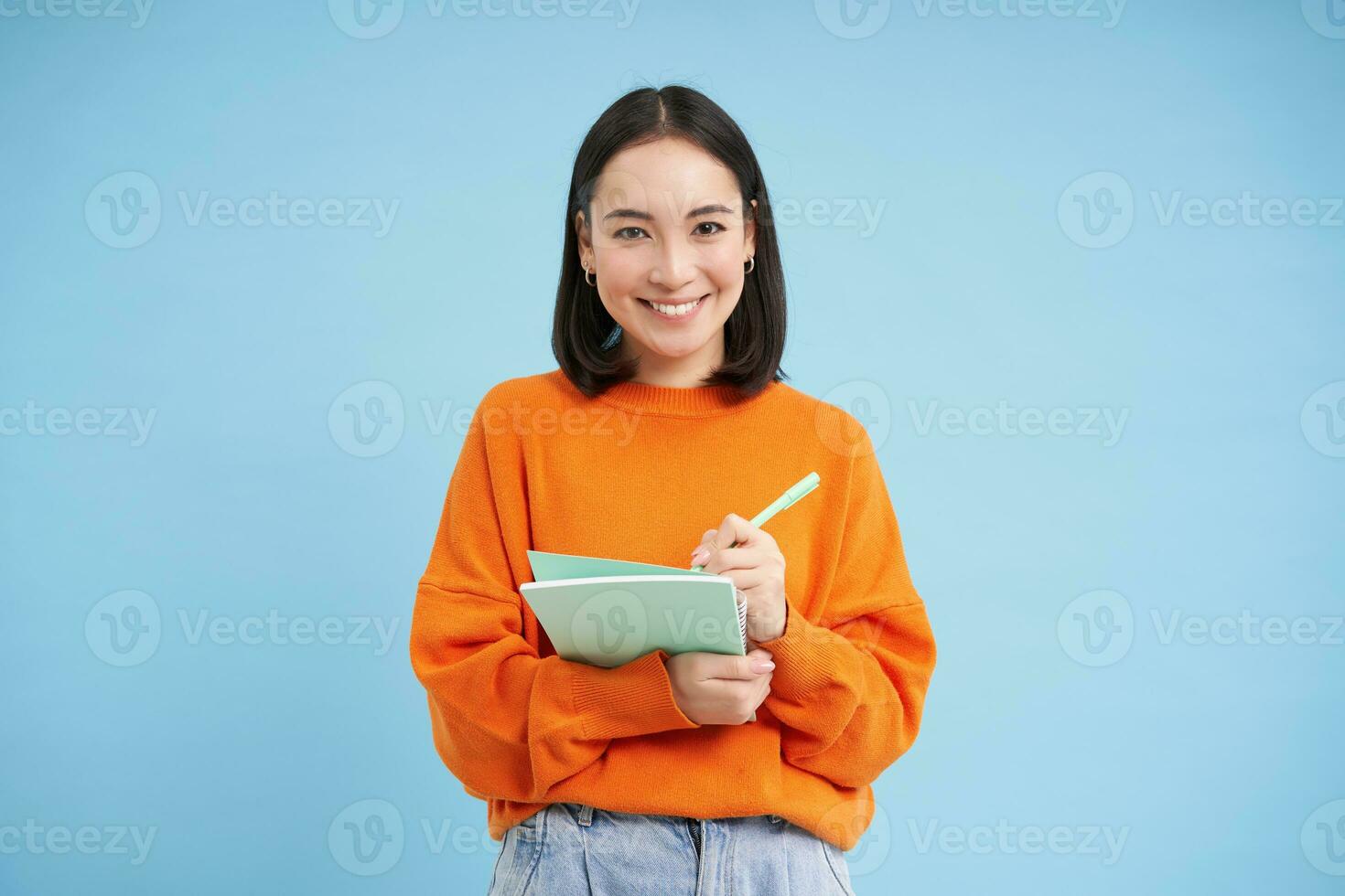Smiling cute girl student, making notes, writing down homework, studying, standing over blue studio background photo