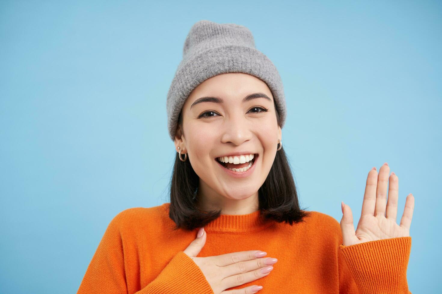 Close up of cute korean girl in beanie, raises one hand and introduces herself, greets you, smiles and looks friendly, blue background photo