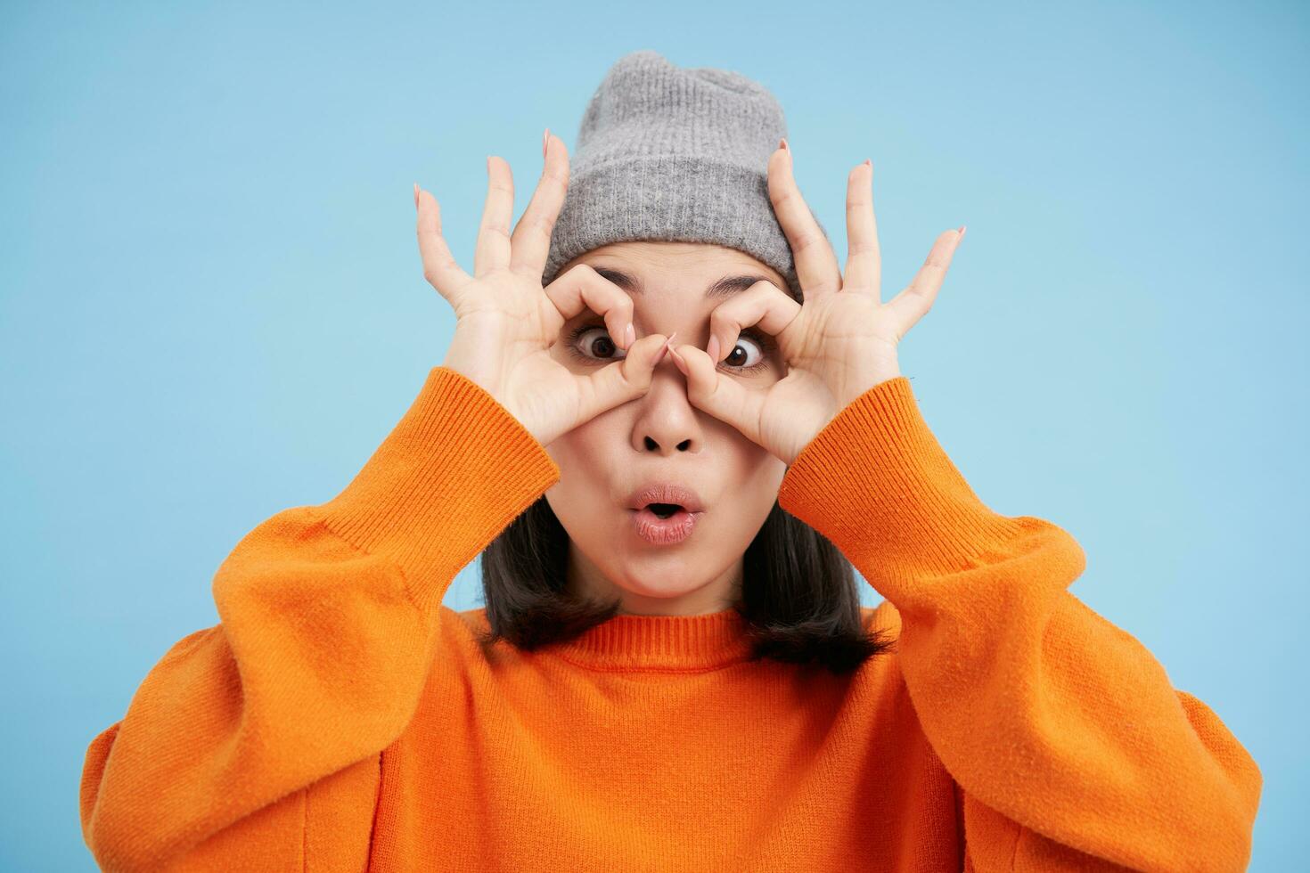 Close up portrait of funny Chinese girl, looks through hand glasses with surprised face expression, standing over blue background photo