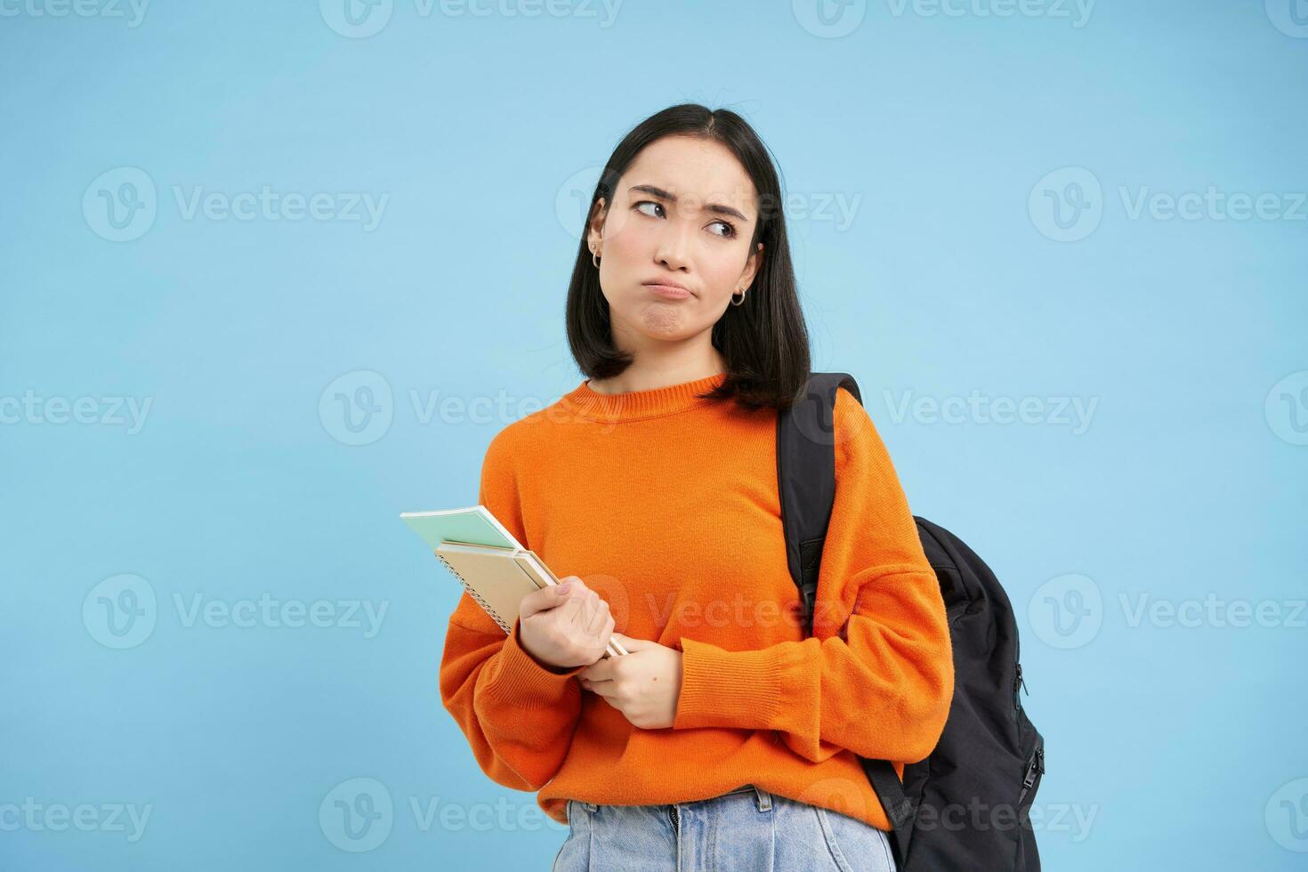 Bored college student, girl with backpack and notebooks, sulks and rolls eyes, stands upset against blue background photo
