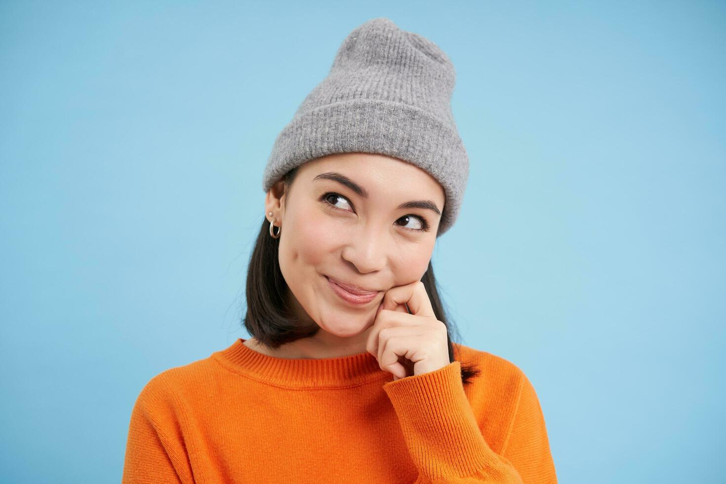 Close up portrait of coquettish asian girl in hat, looks aside and smiling, fantasizing, dreaming of smth, standing over blue background photo