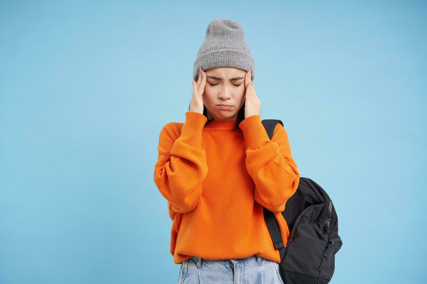 Distressed asian woman holds hands on head, looks worried and concerned, stands over blue studio background photo