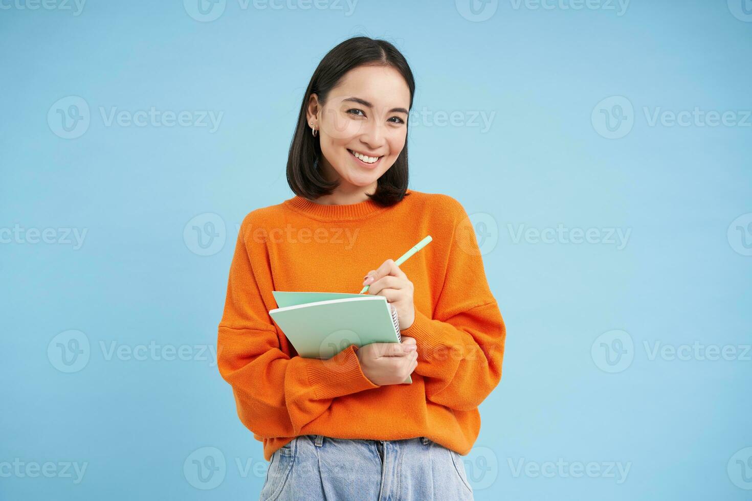 Education and students. Happy asian woman, holding notebooks and laughing, smiling at camera, enjoys going to University or College, blue background photo