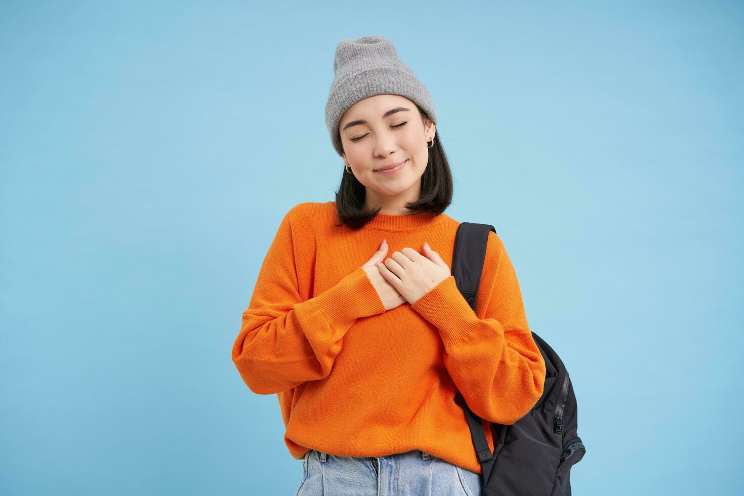 Romantic young woman in hat, holds backpack on shoulder, holds hands on heart and closes eyes, dreaming of something, standing over blue background photo