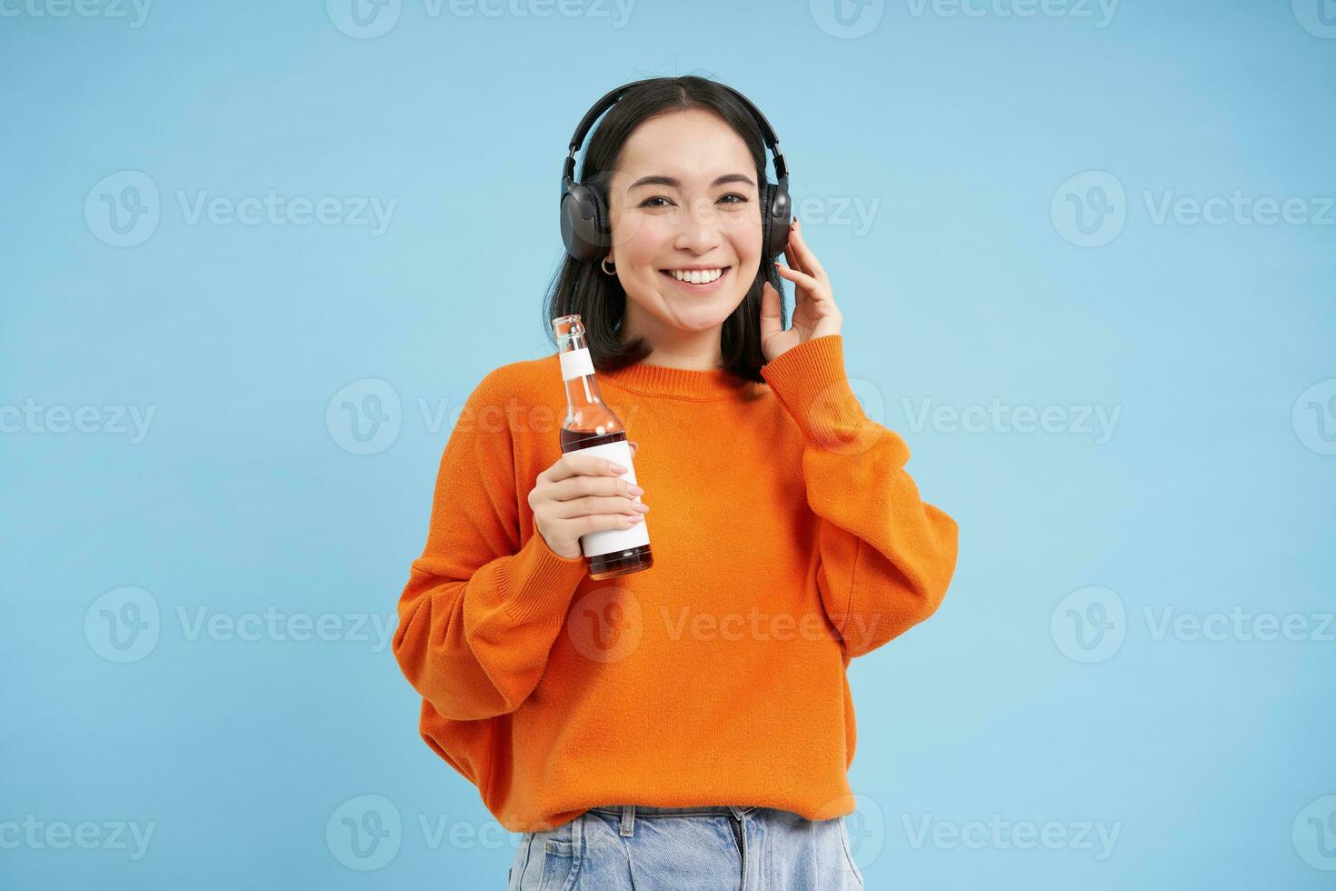 Beautiful asian woman listens music in headphones, drinks coke from bottle and smiles at camera, blue background photo