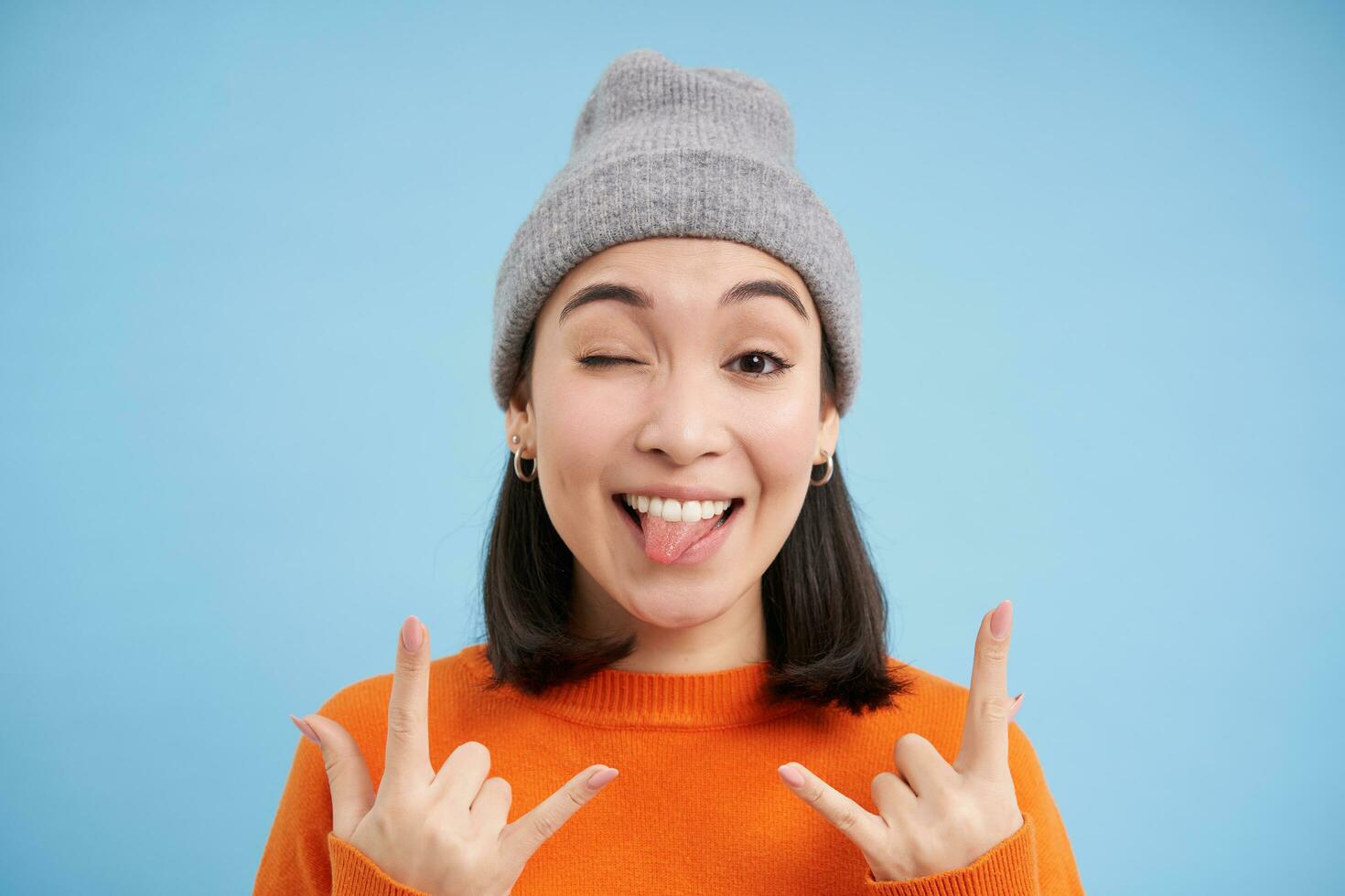 cerca arriba retrato de gracioso y despreocupado asiático niña en gorro, muestra rock norte rollo, pesado metal gesto y lengua, soportes terminado azul antecedentes foto