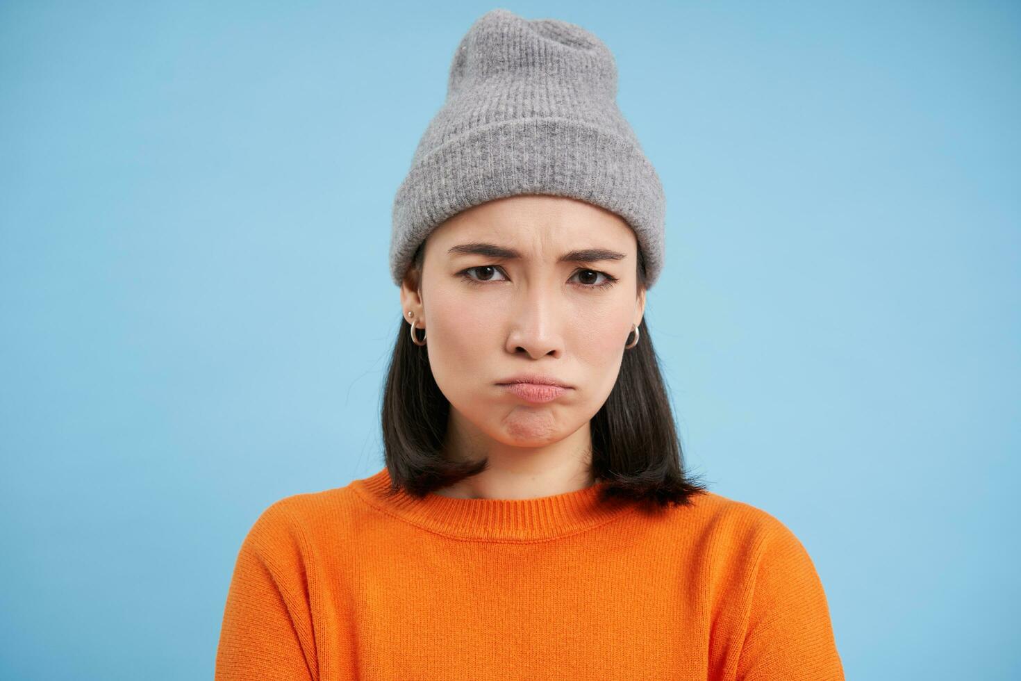 Close up portrait of sulking, sad asian woman in winter hat, frowning and looking angry, offended face, standing over blue background photo