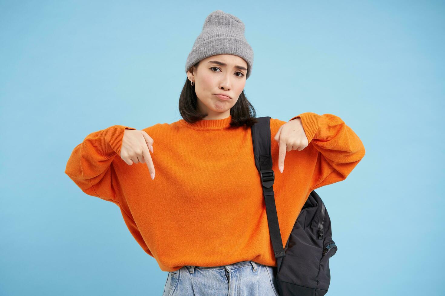 Asian woman in warm hat, wears street outfit and backpack, points fingers down, shows advertisement below, standing over blue background photo