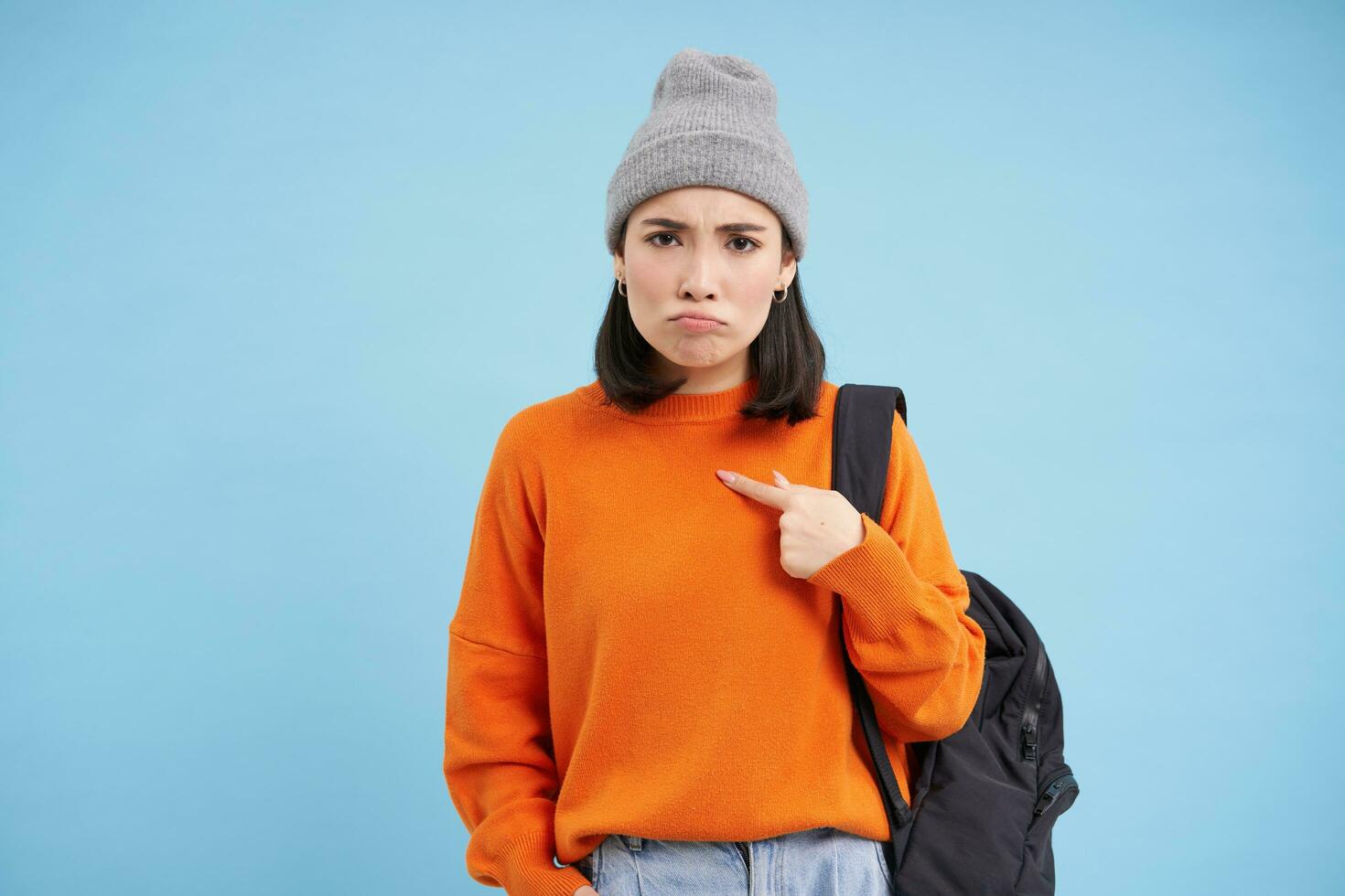 Sad asian girl points at herself with upset, gloomy face expression, stands over blue studio background photo