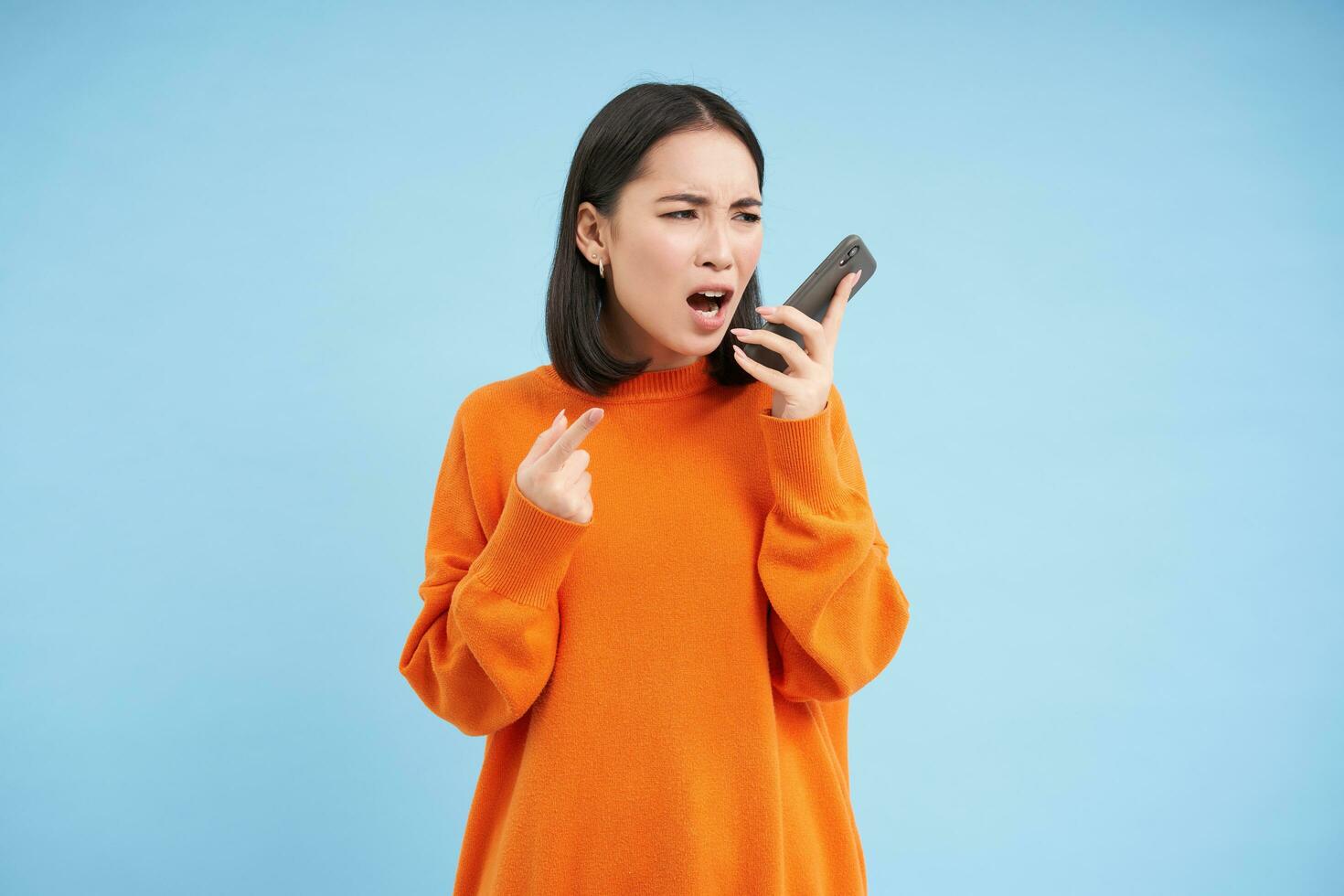 Angry asian woman yells at mobile phone, shouts in smartphone with furious face, stands over blue background photo