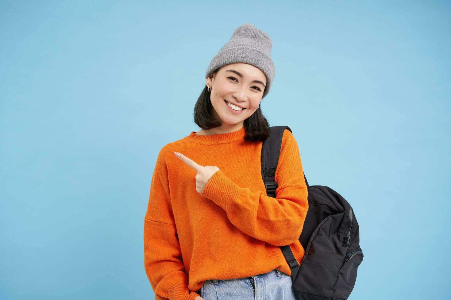 Enthusiastic asian woman, points finger left at banner, shows promo offer, smiles and looks happy, stands in hat with backpack, blue background photo