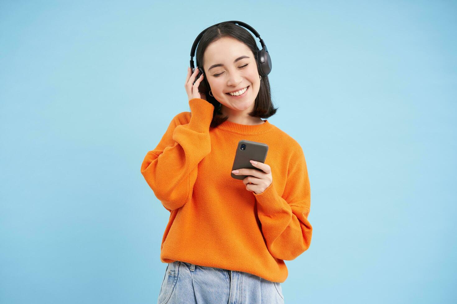 Happy young modern woman with headphones and smartphone, listening to music on mobile phone, enjoys favourite podcast, blue background photo