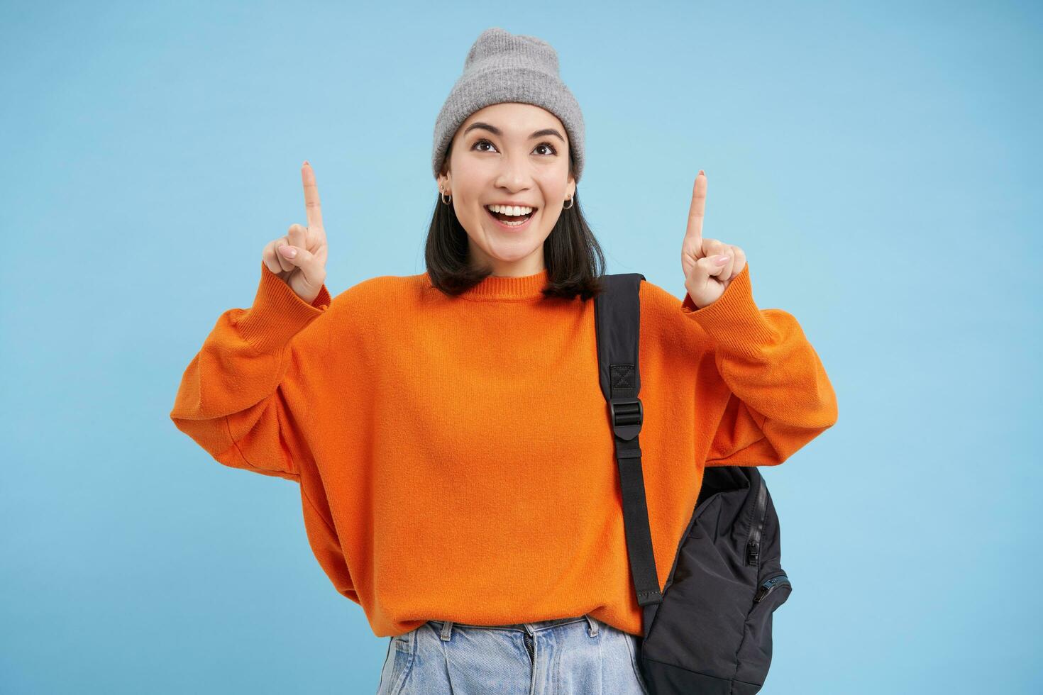 Happy korean girl points fingers up, shows advertisement, invites to click banner on top, stands over blue background photo