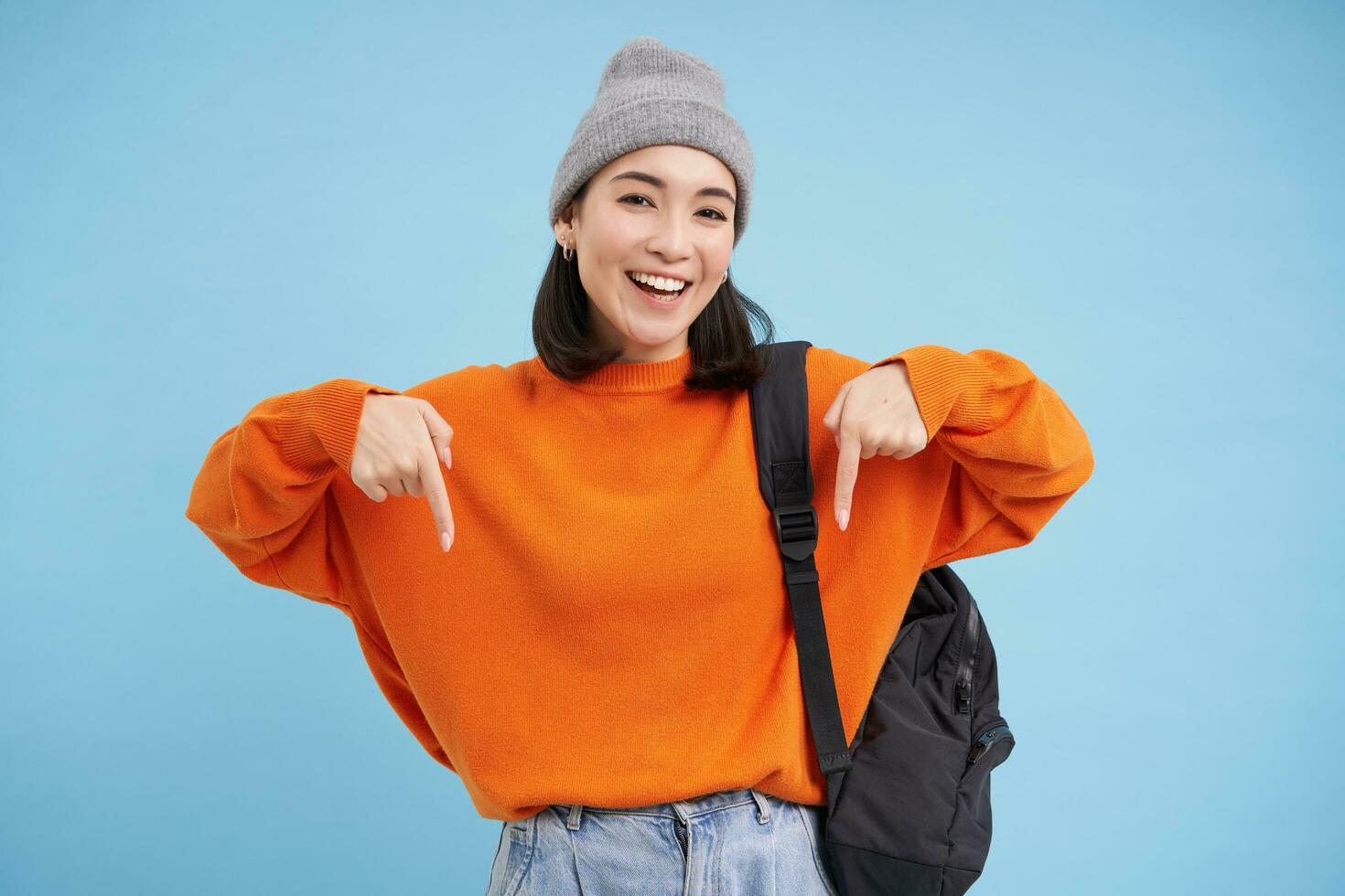 Asian woman in warm hat, wears street outfit and backpack, points fingers down, shows advertisement below, standing over blue background photo