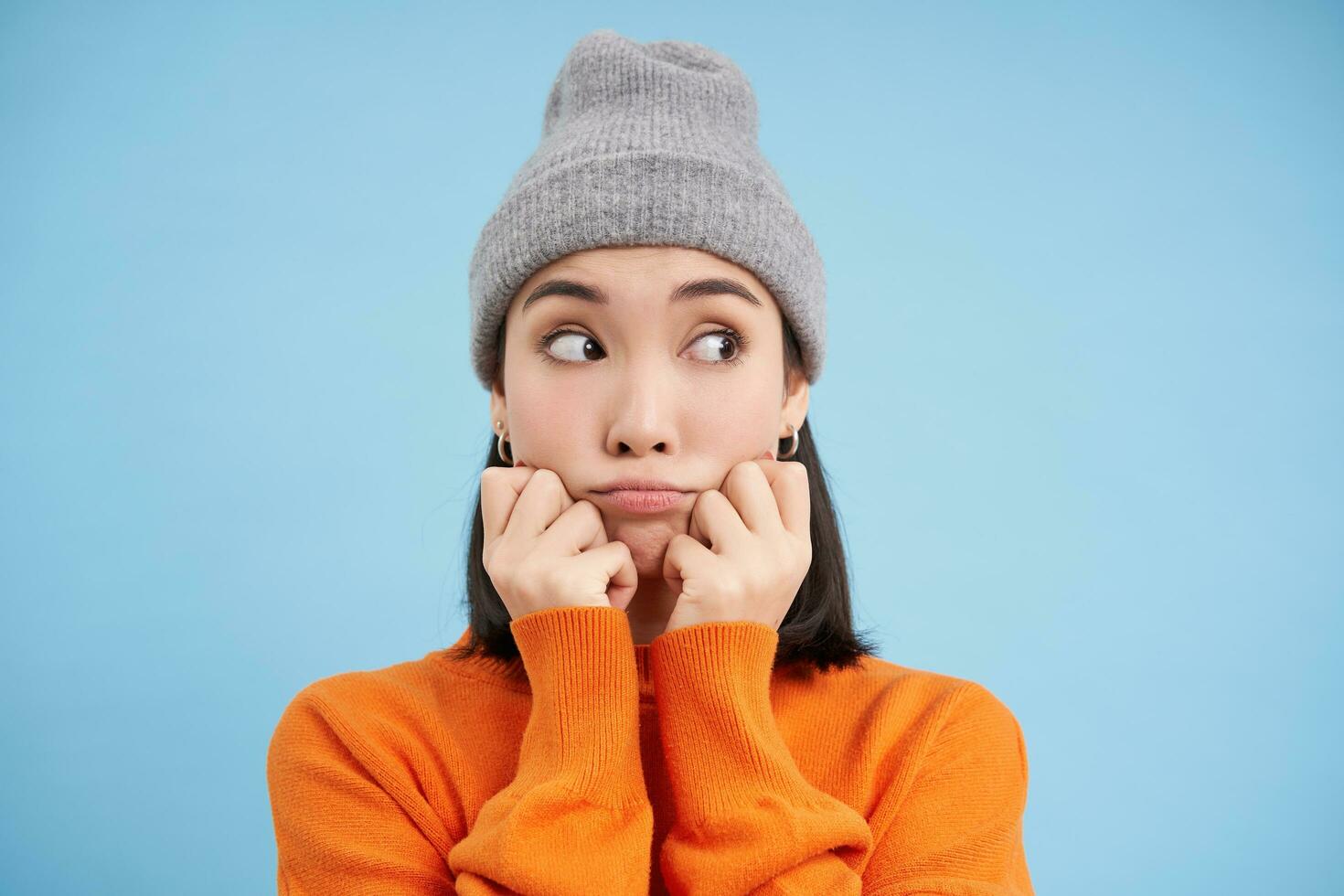 Close up portrait of cute korean girl in warm beanie, pouting and looking cute aside at blank copy space on blue background photo