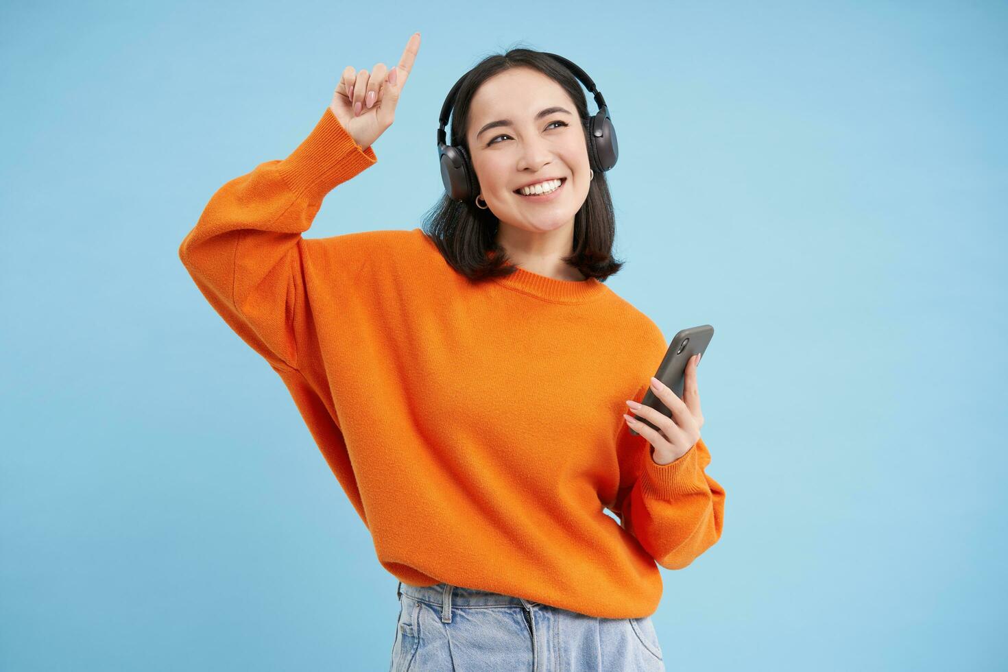 Beautiful korean woman in headphones, dancing, listening music on mobile phone app, smiling, posing over blue background photo
