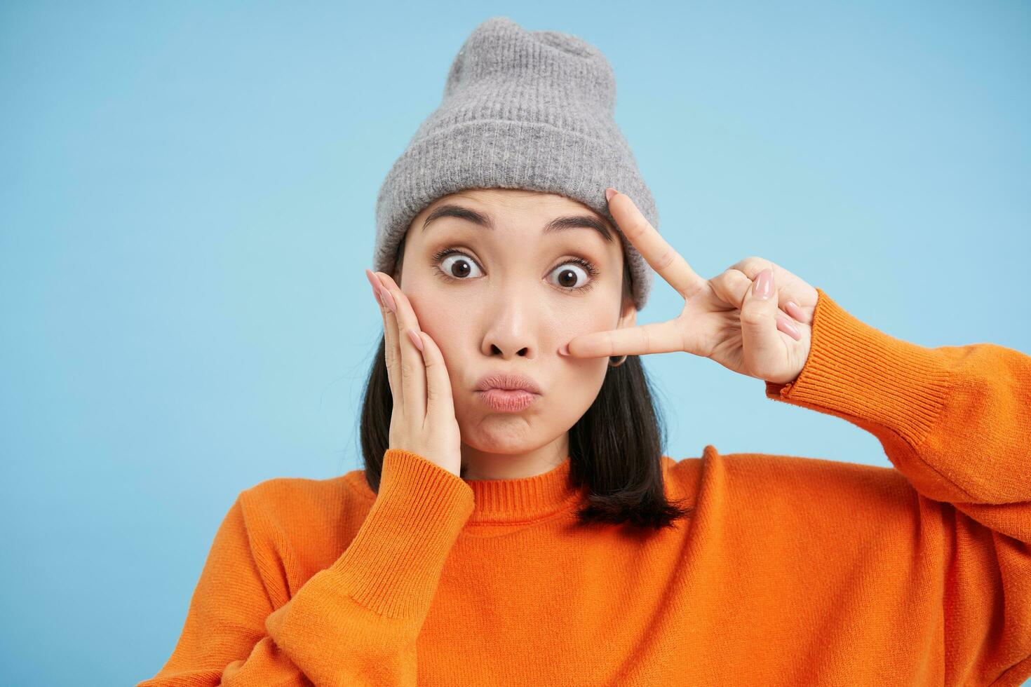 cerca arriba retrato de linda niña en gorro, cubos labios y muestra paz firmar, posando kawaii en contra azul antecedentes foto