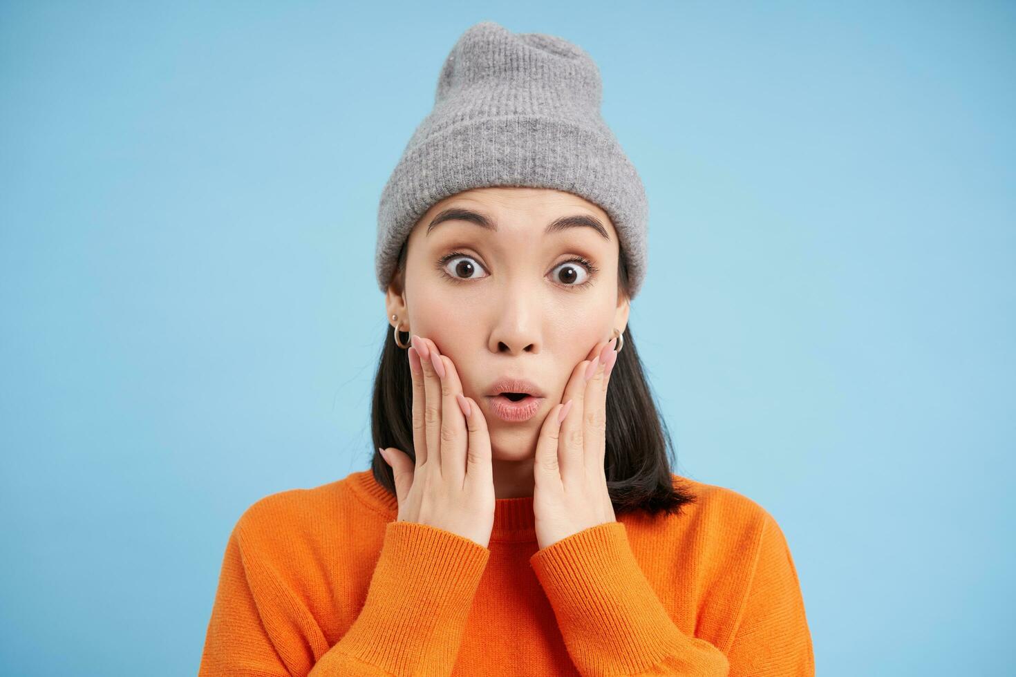 Surprised korean girl in warm hat, looks amazed at camera, expresses interest and amazement, stands in orange sweatshirt over blue background photo