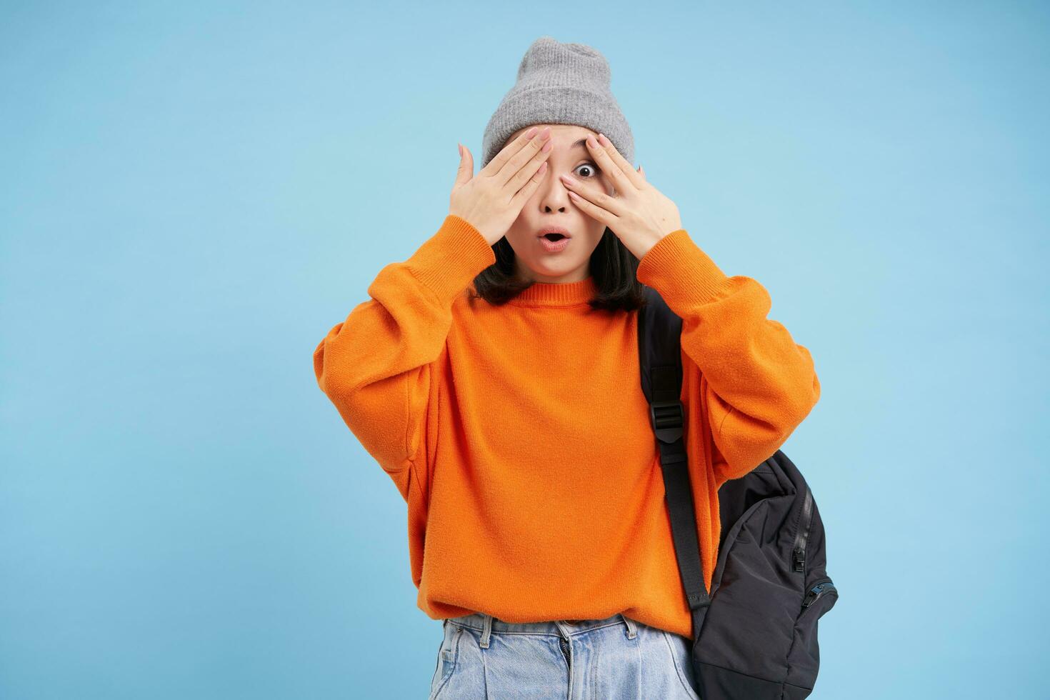 Cute asian woman with backpack, closes eyes and waits for surprise, stands over blue background photo