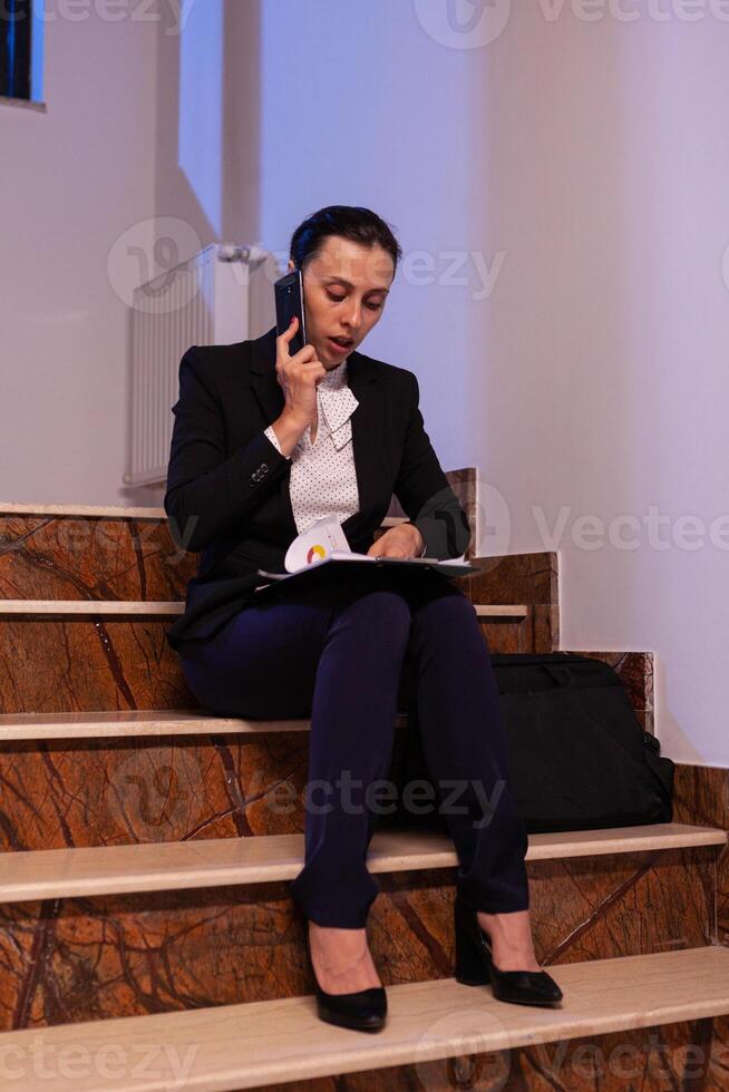 Overworked businessoman disccusing about project deadling on smartphone late at night doring overtime. Serious entrepreneur working on job project sitting on staircase of business building at night hours for job. photo