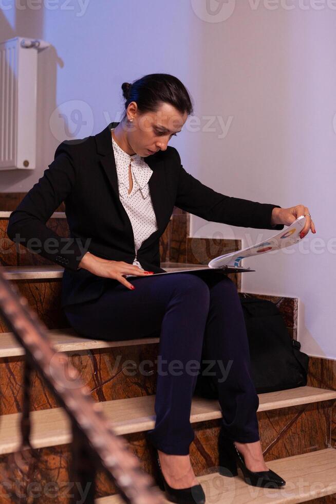 Exhausted tired overworked businesswoman holding financial reaport of deadline project. Serious entrepreneur working on job project sitting on staircase of business building at night hours for job. photo
