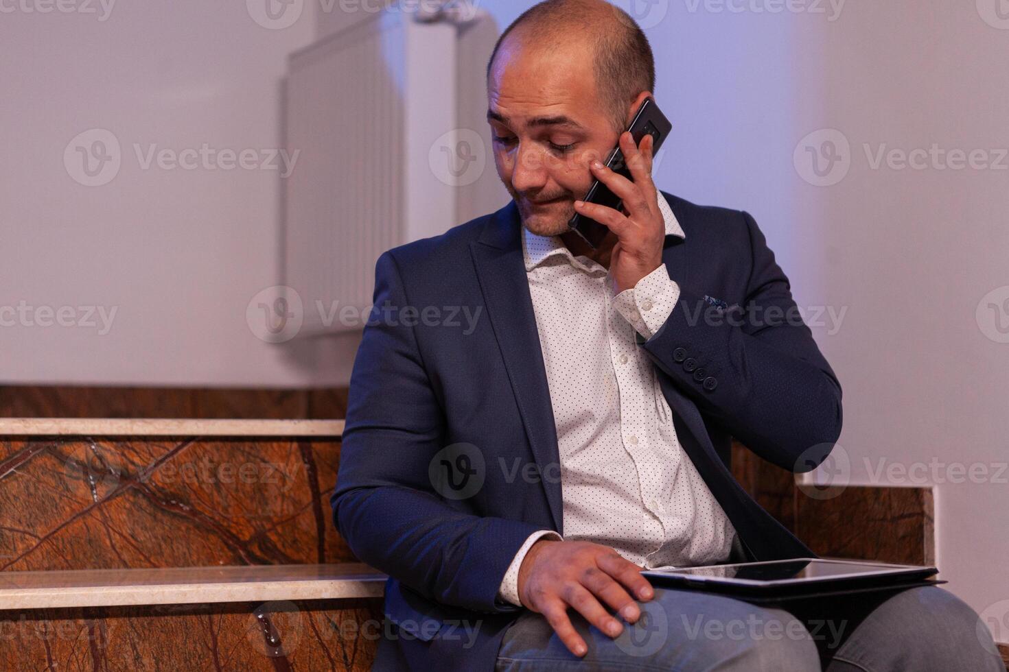 Exhausted overworked businessman talking on phone with team about project deadline.Entrepreneur working late in evening corporate sitting on stairs in building office. photo