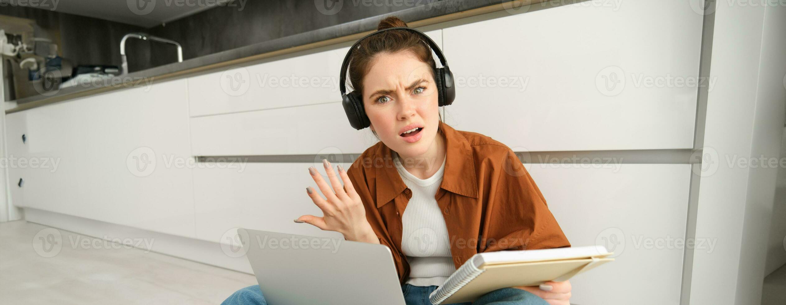 Portrait of annoyed and confused woman in headphones, sits on floor with laptop and notebook, looks frustrated, has issues in doing homework or studying photo