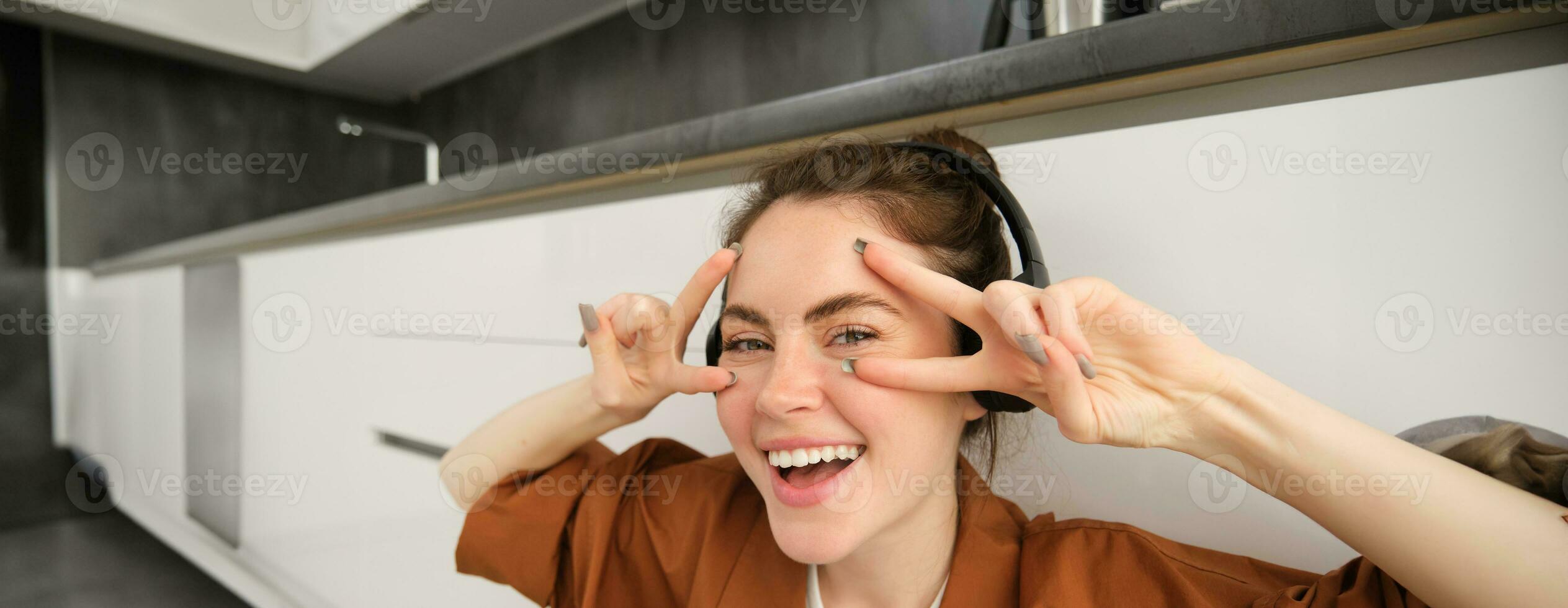 Close up of carefree young woman, laughing and smiling, showing peace, v-sign gesture, listening to music in wireless headphones photo