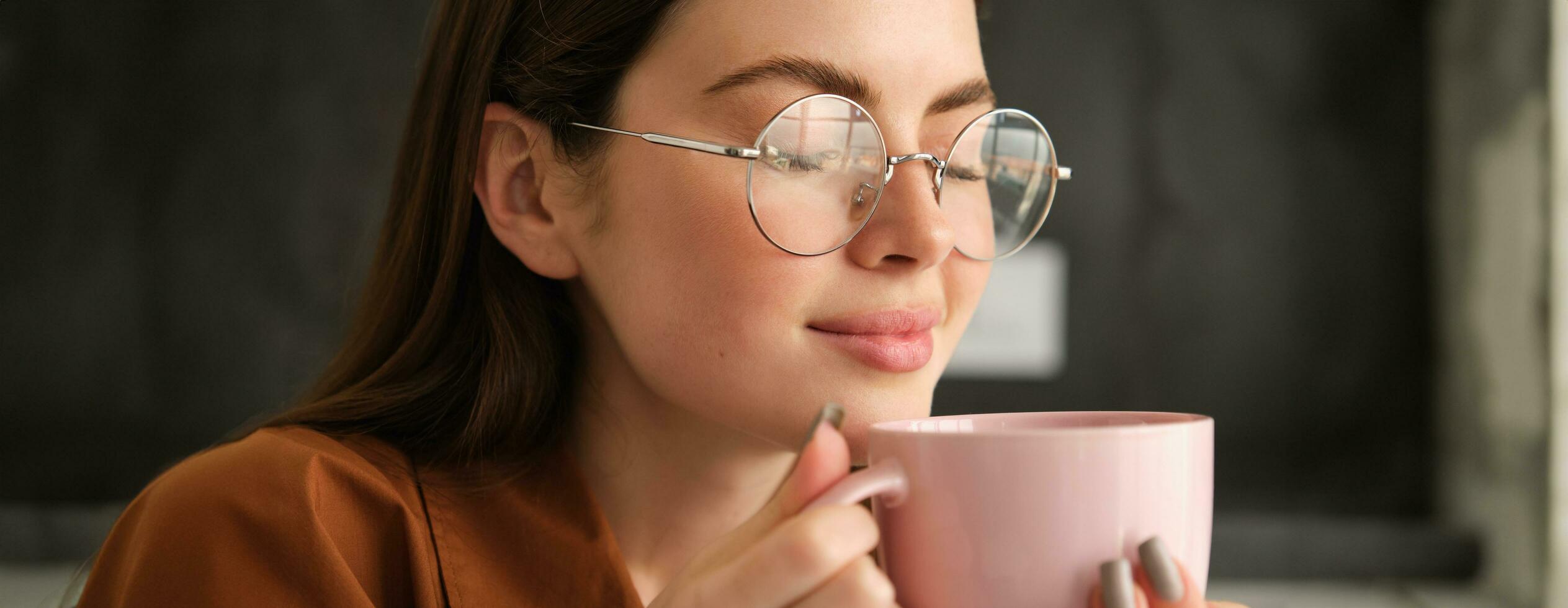 cerca arriba retrato de hermosa sonriente mujer, bebidas su Mañana taza de café, disfruta sabroso aroma, oliendo su bebida con satisfecho cara foto