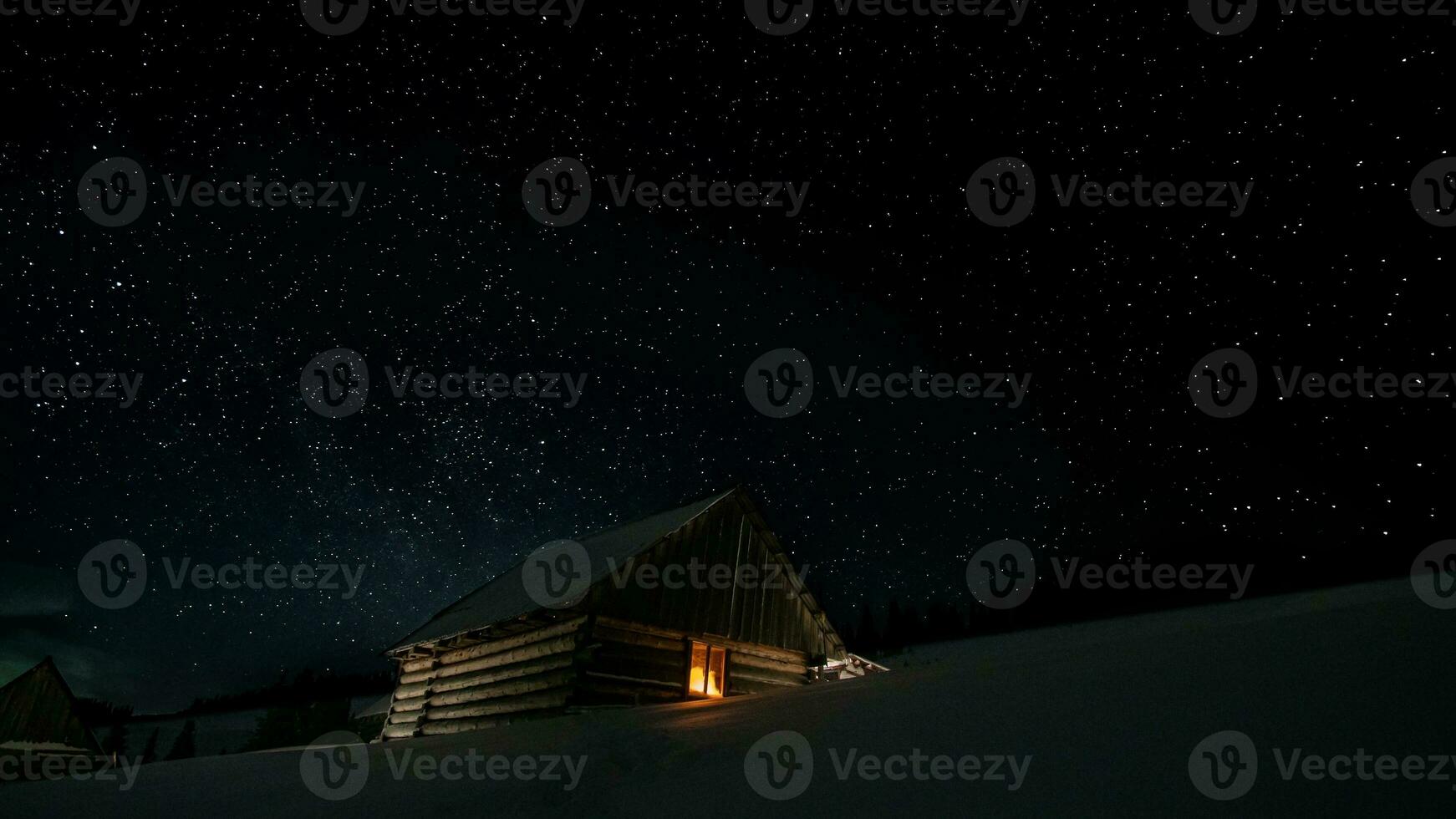 Stars in the night sky and a wooden house with a glowing window in winter photo