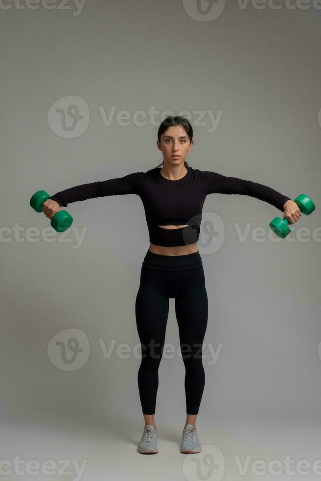 Sporty girl performing dumbbell lateral raise on grey background photo