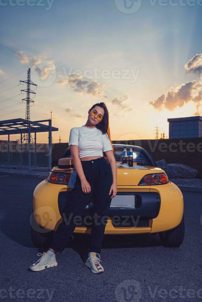 Woman in sunglasses, white top, black pants and sneakers is posing outdoors near yellow cabriolet with two bottles of soda on trunk. Full length photo