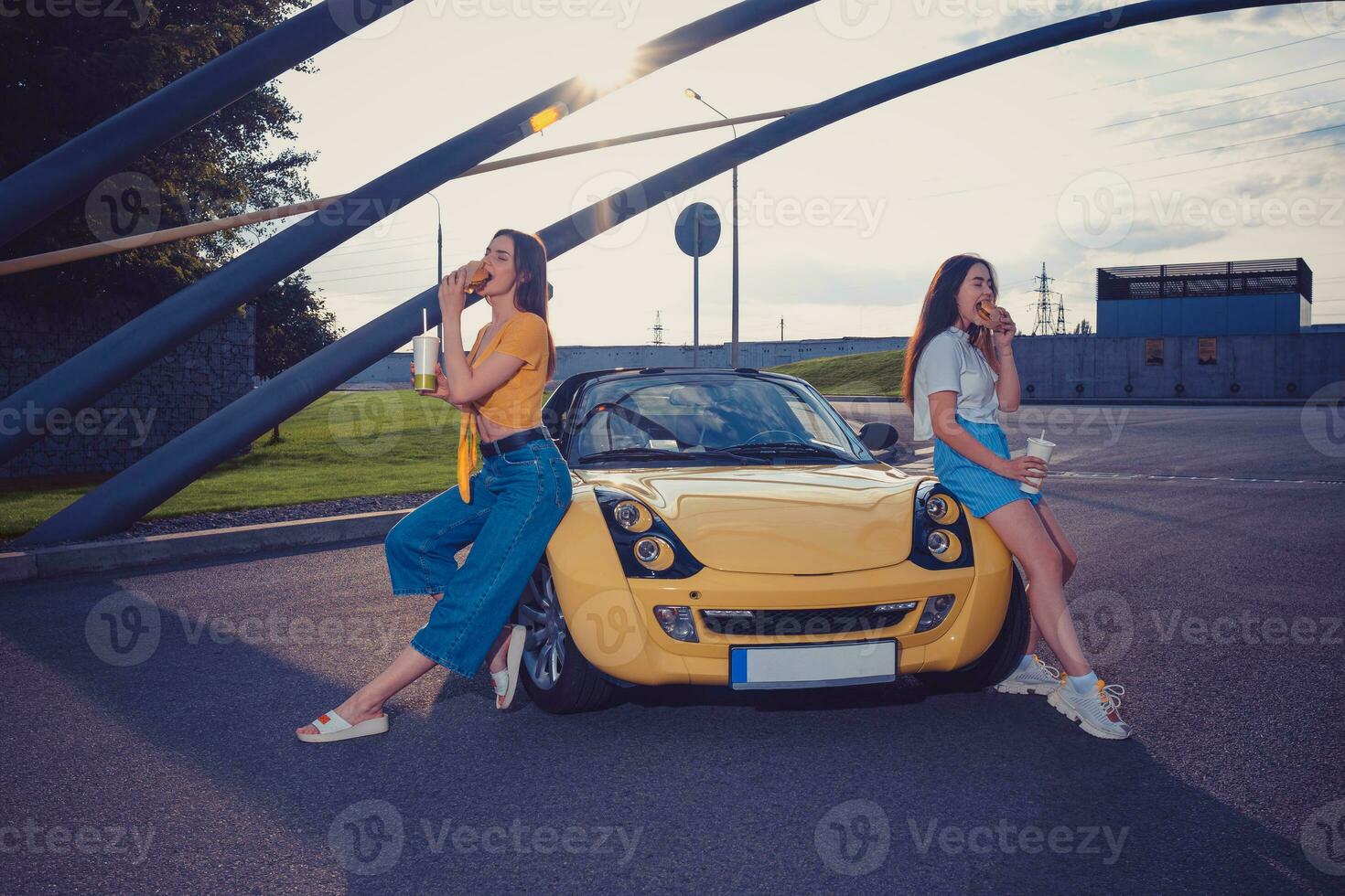 joven modelos son comiendo hamburguesas y participación bebidas en papel tazas mientras propensión en el capucha de amarillo coche. rápido alimento. Copiar espacio, burlarse de arriba foto