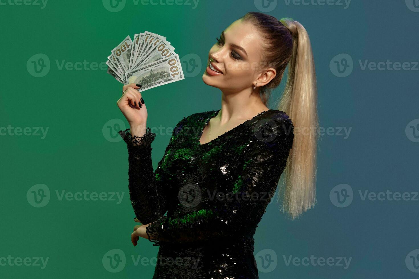 Blonde girl in black sequin dress. Smiling, propping her elbow with hand, showing some money, posing on colorful background. Poker, casino. Close-up photo