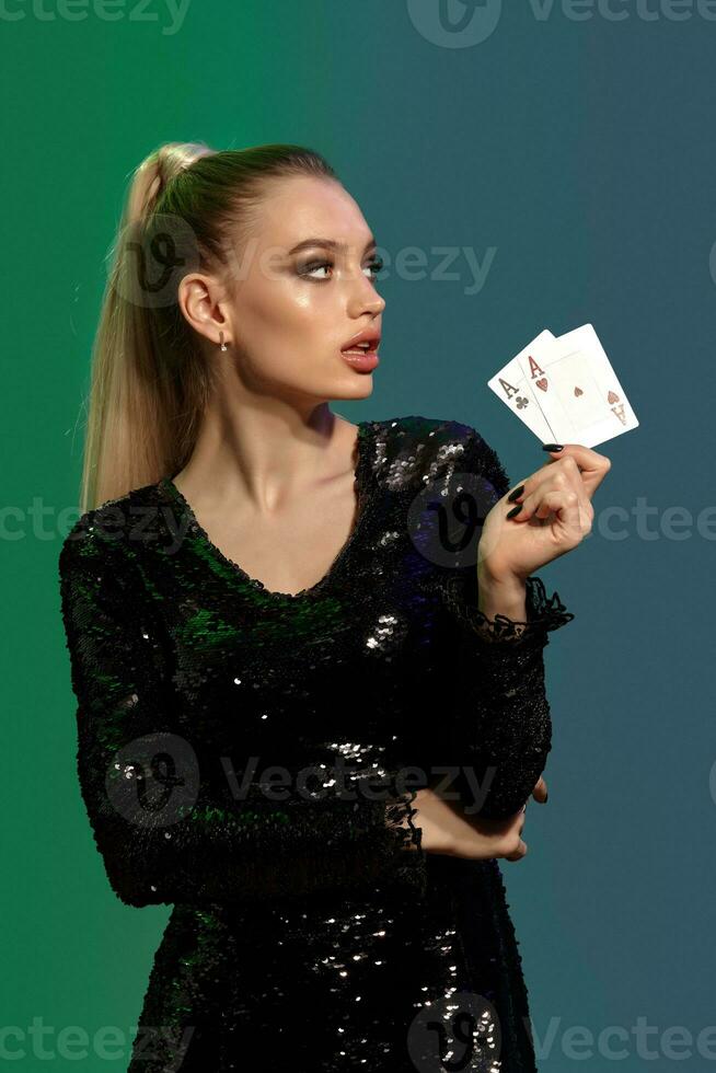 Blonde model in jewelry and black sequin dress. She is showing two playing cards, posing on colorful background. Gambling, poker, casino. Close-up photo