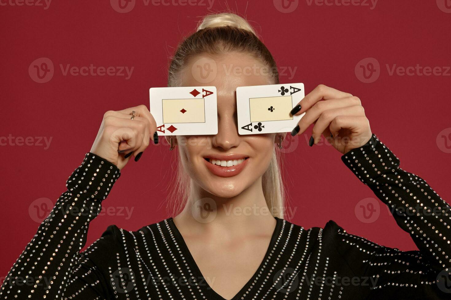 Blonde girl in black dress in rhinestones. Smiling, closed her eyes with two playing cards, posing on red background. Poker, casino. Close-up photo